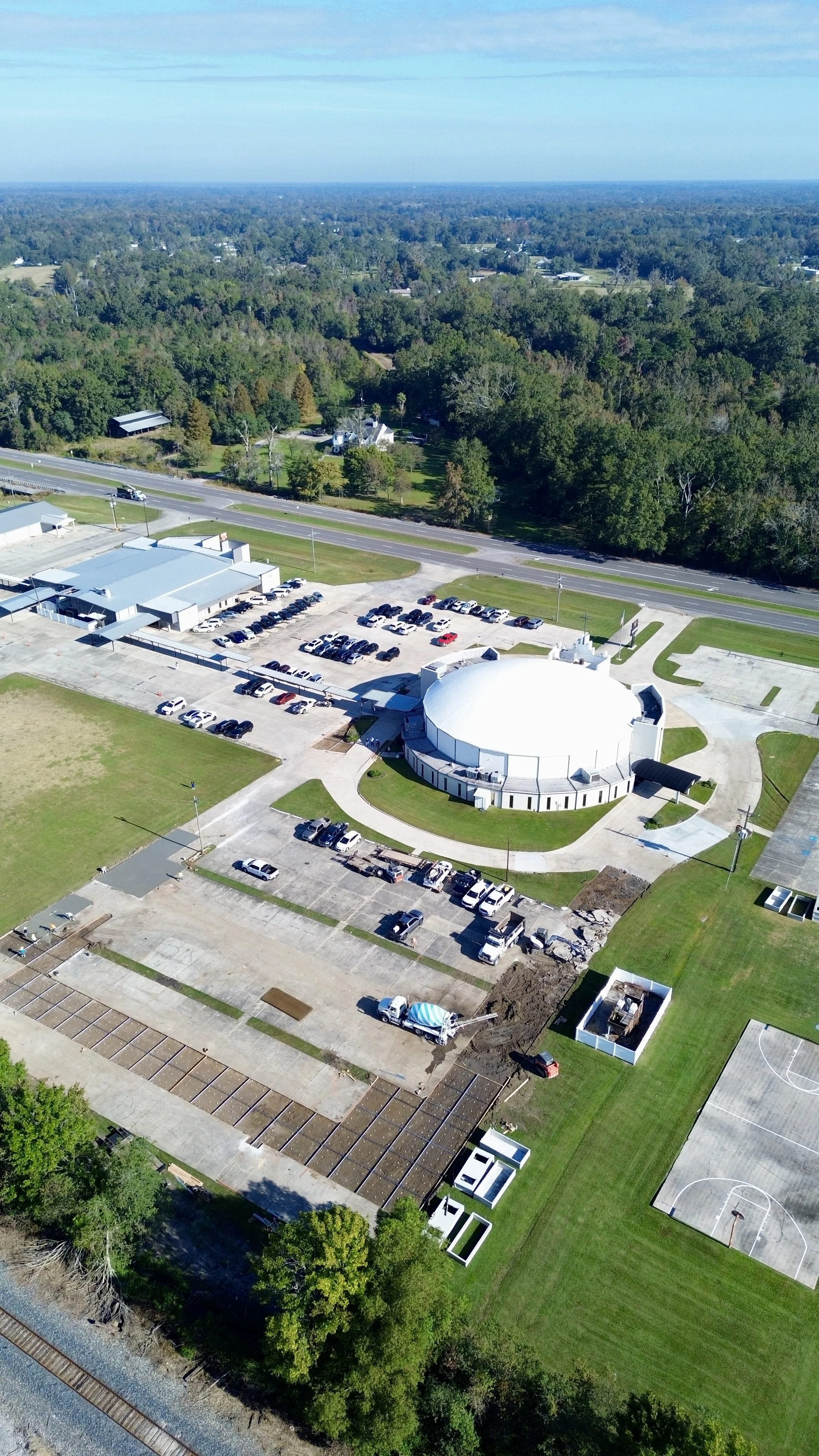 An aerial view of a building with a white dome roof, parking lot with cars, sports courts, and surrounding greenery.