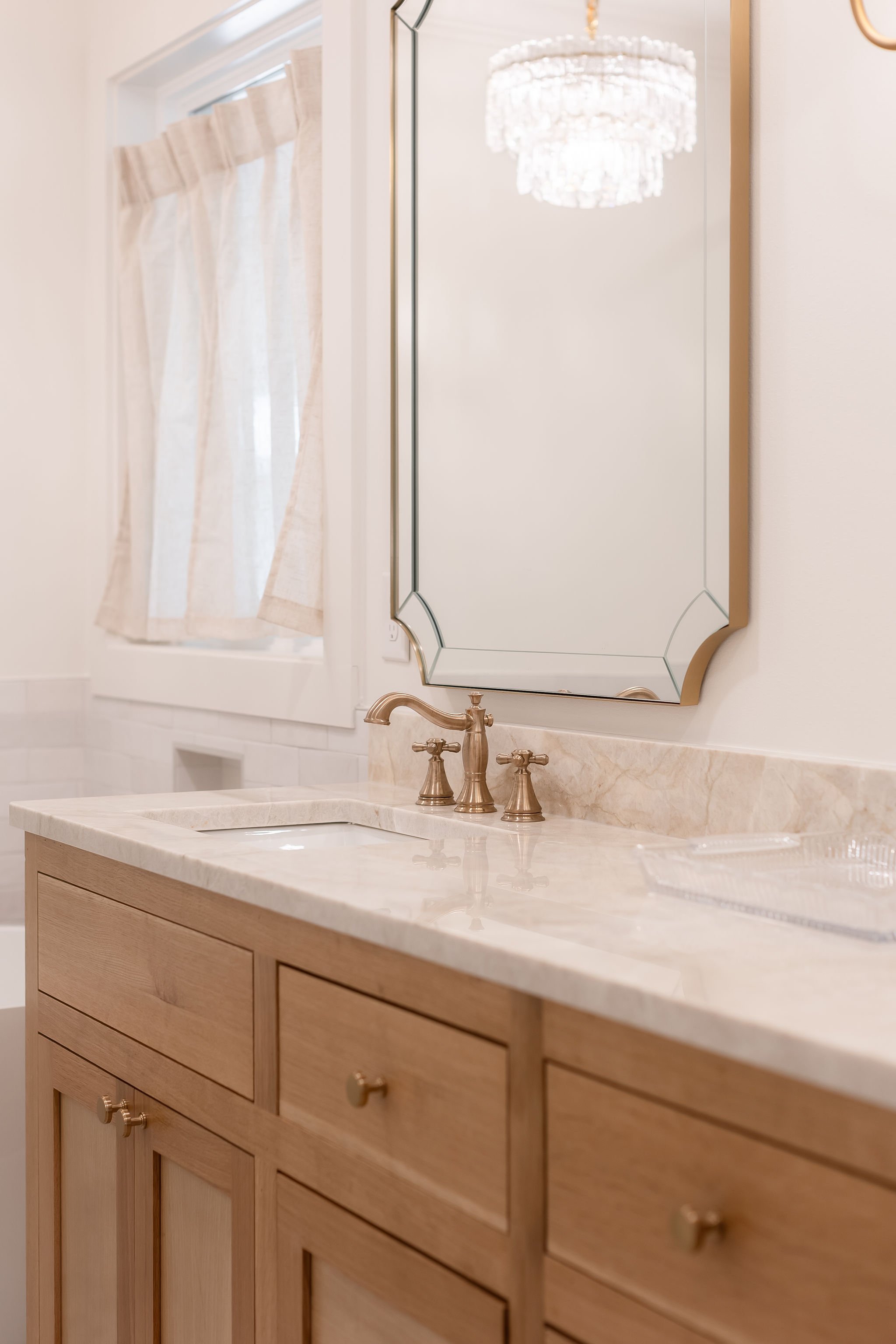 Bathroom vanity with a marble countertop, a mirror, and brass fixtures, with a window and chandelier reflected in the mirror.