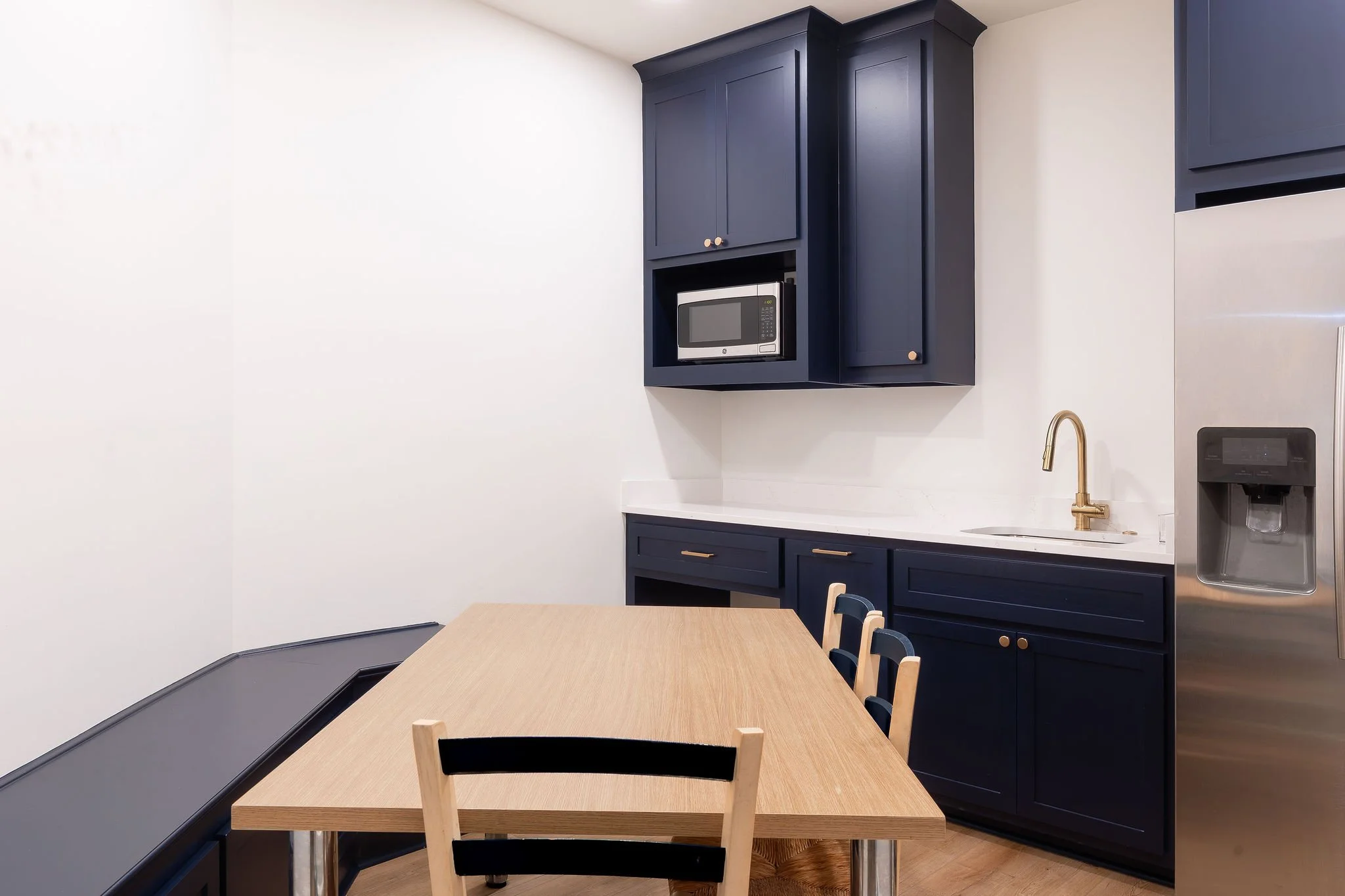 Modern kitchen with blue cabinets, a microwave oven, a stainless steel refrigerator, a white marble countertop, a gold faucet, and a wooden dining table with chairs.