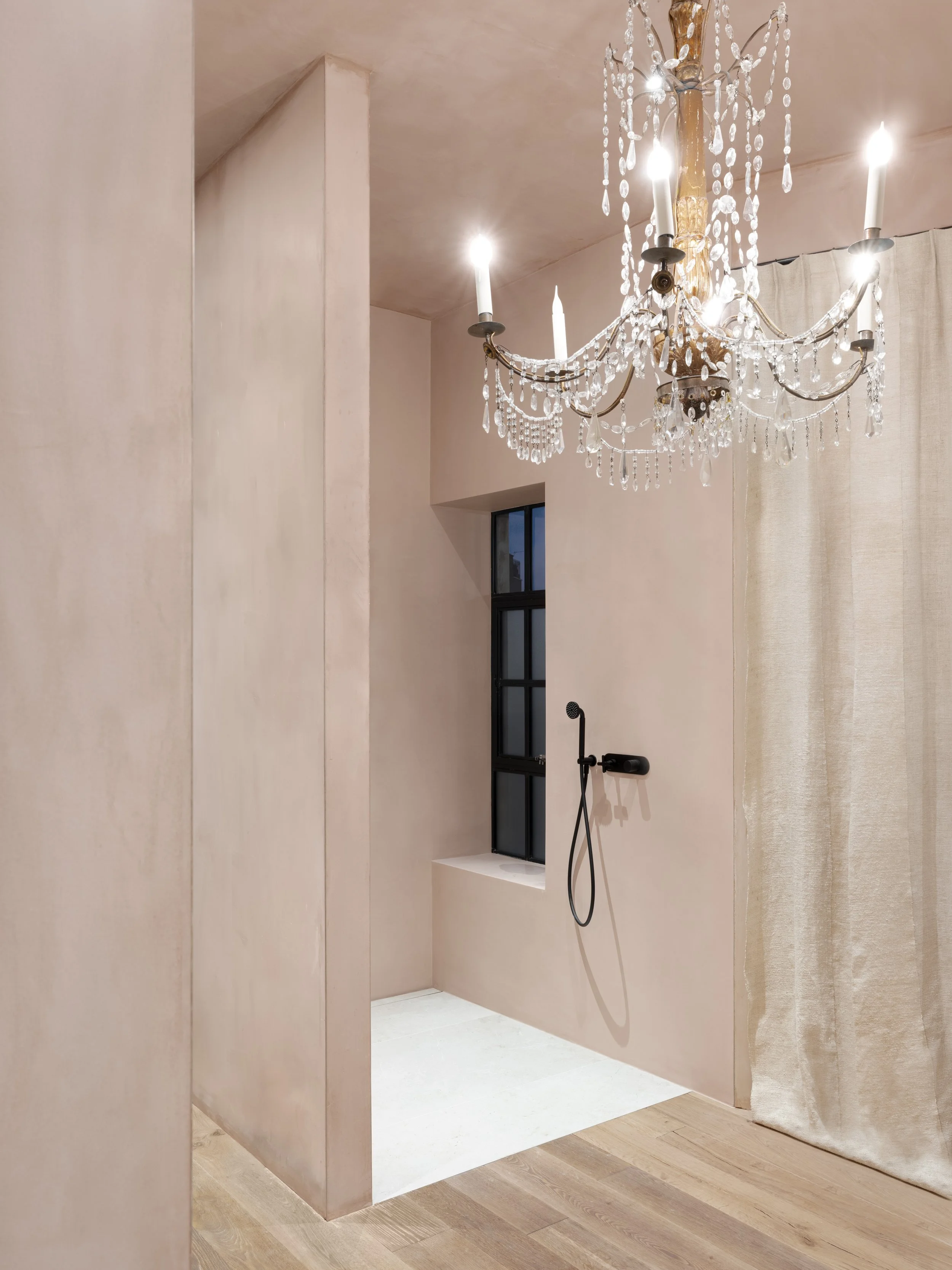 A luxurious bathroom with a black showerhead on a blush pink wall, a small window, a chandelier with crystal accents, and beige curtains.