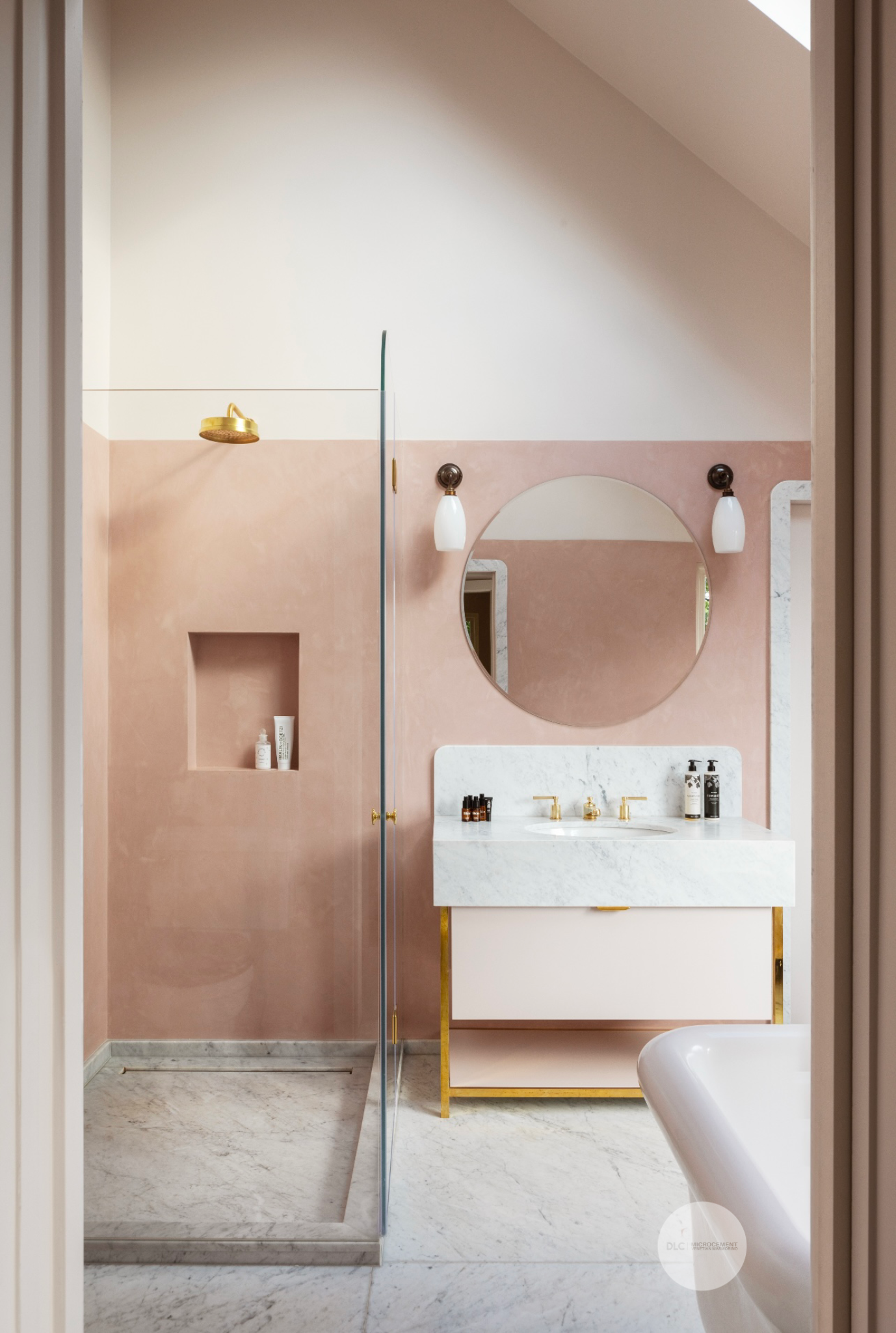 Modern bathroom with a pink wall, round mirror, white marble sink with gold fixtures, and a glass shower with marble base.