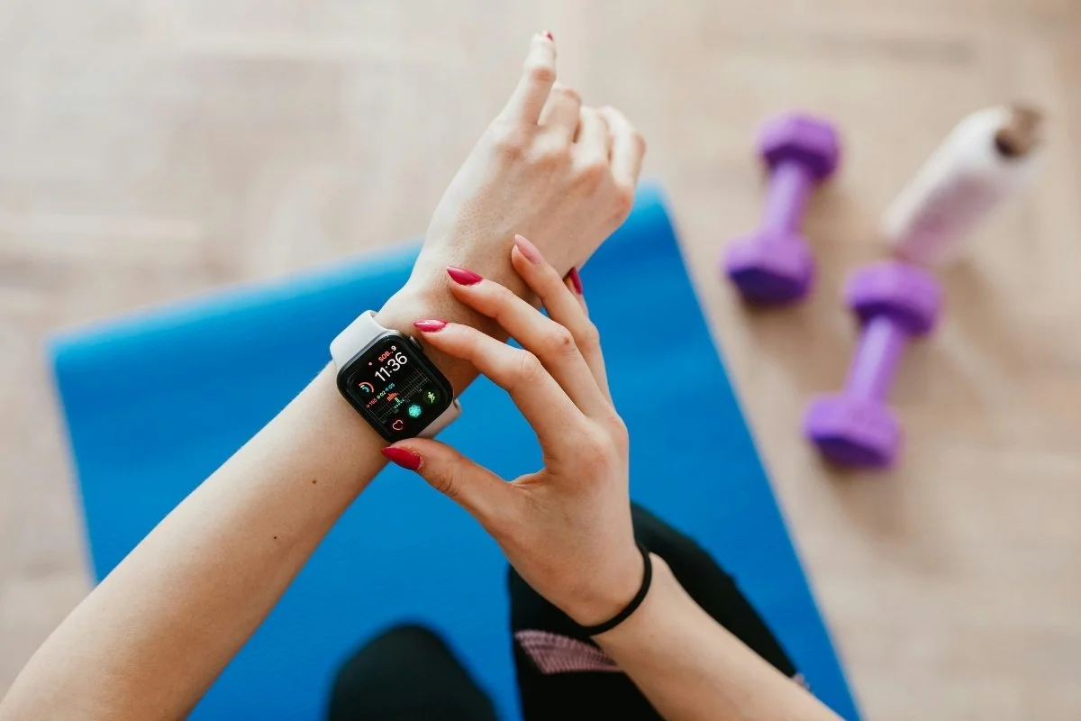 a photo of a young woman's hands as you clicks on a button on her watch with exercise tracking. Below here is a mat and purple bar bells
