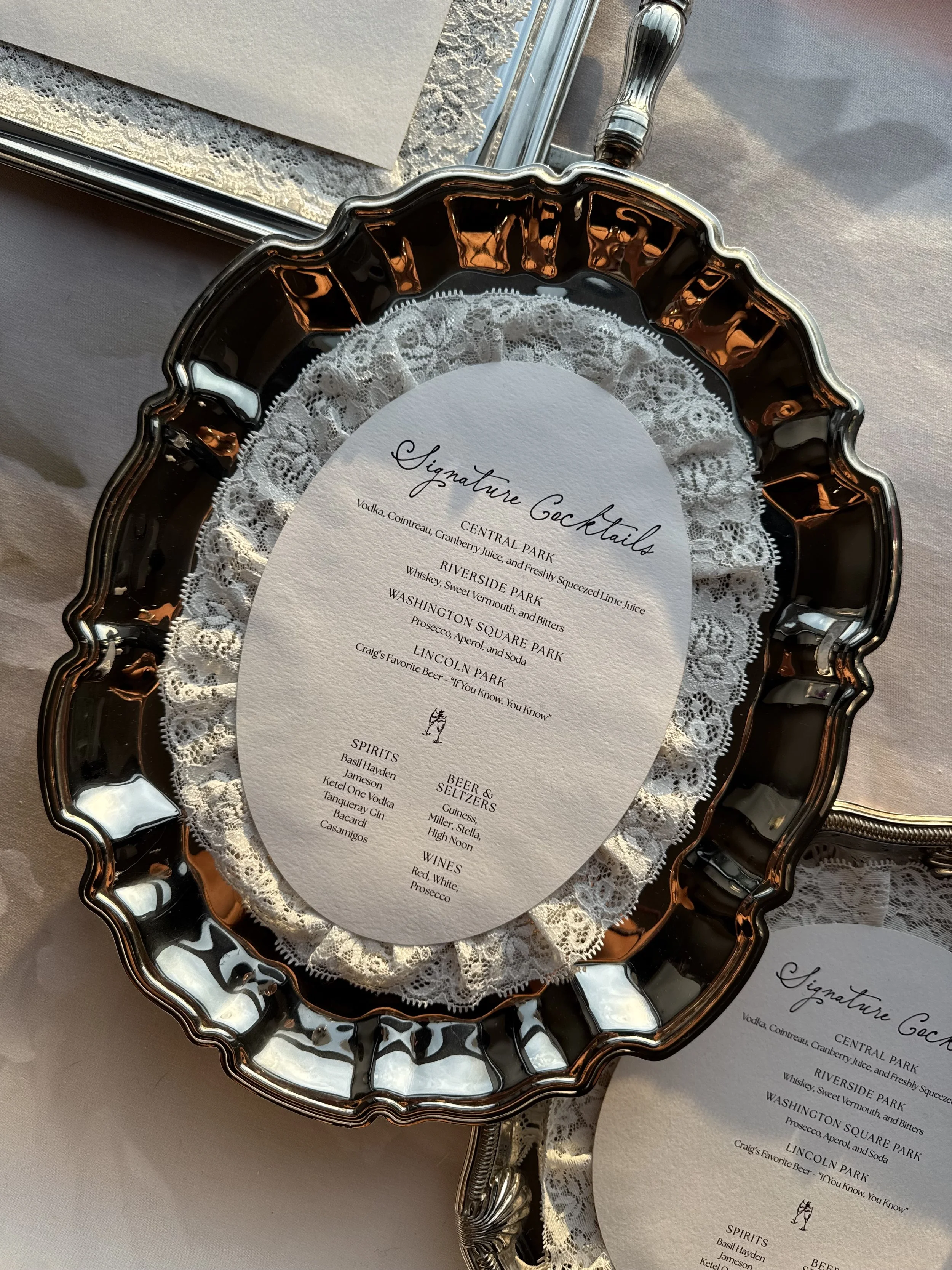 Custom wedding cocktail bar menus displayed on vintage silver platters with delicate lace, oval textured paper, and elegant calligraphy for a romantic wedding reception.