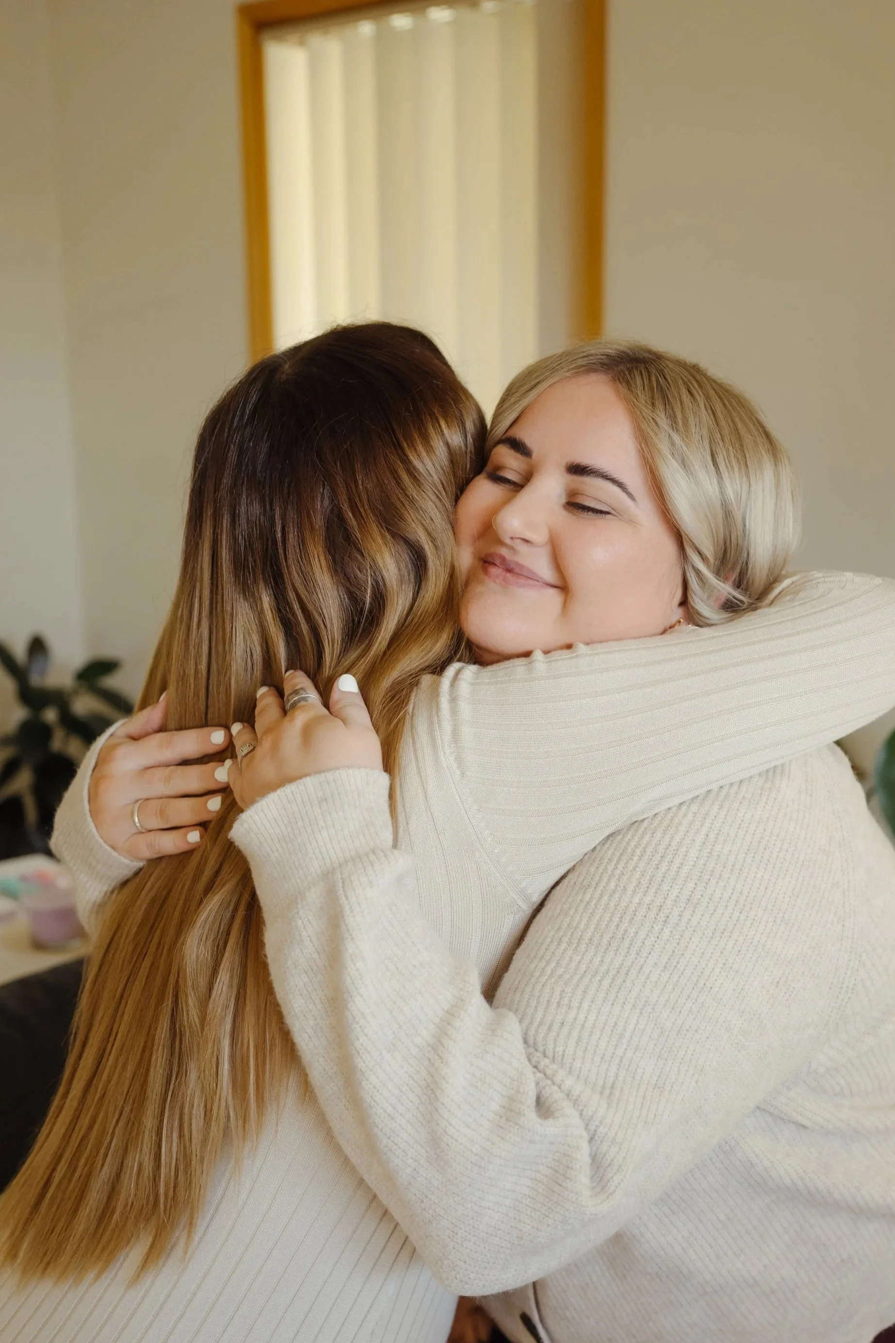 Two women hugging warmly, with one woman smiling and eyes closed, in a cozy indoor setting.