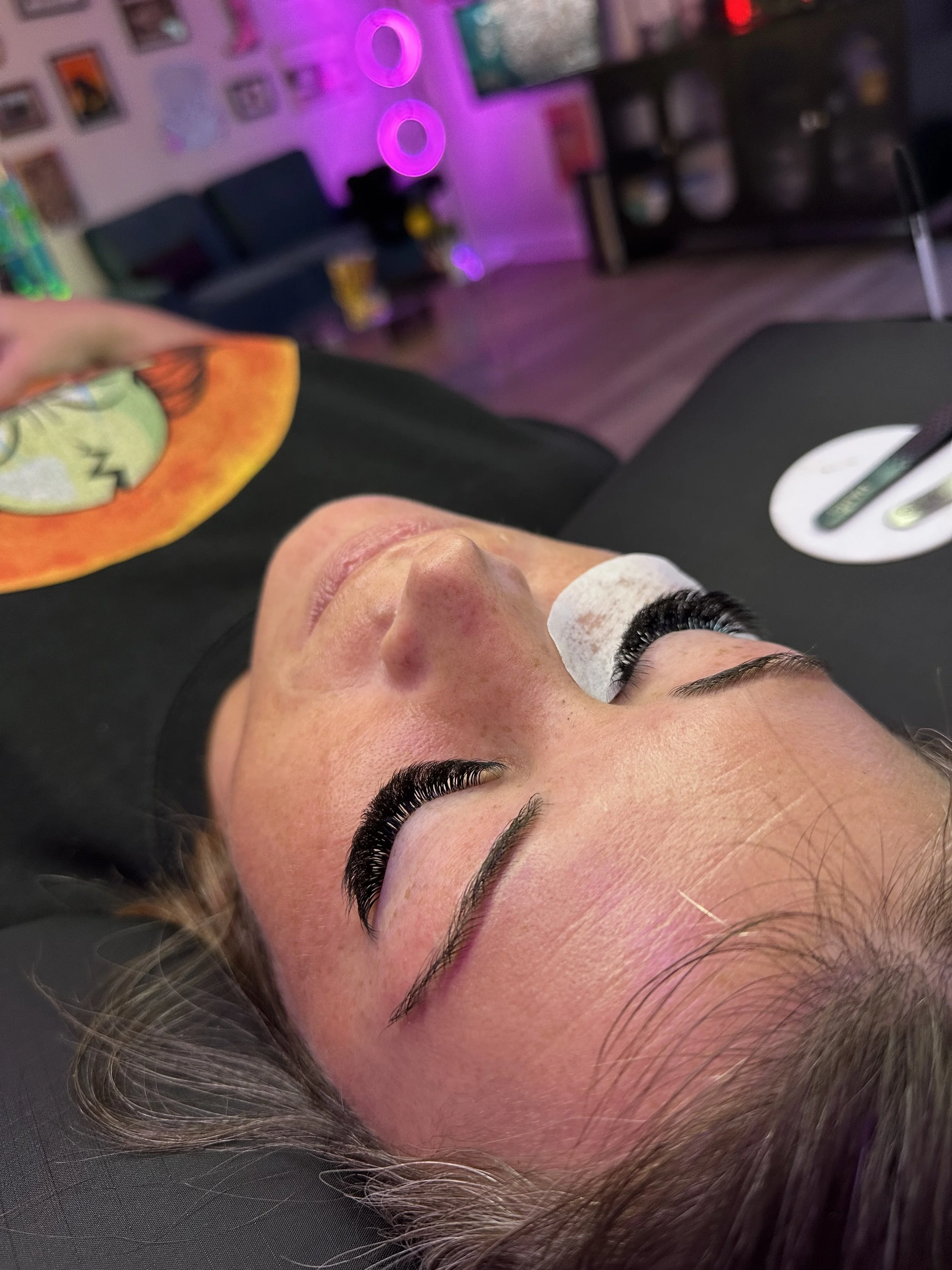 A woman lying down, receiving eyelash extensions, with her eyes closed in a beauty salon with pink and purple colored lights in the background.