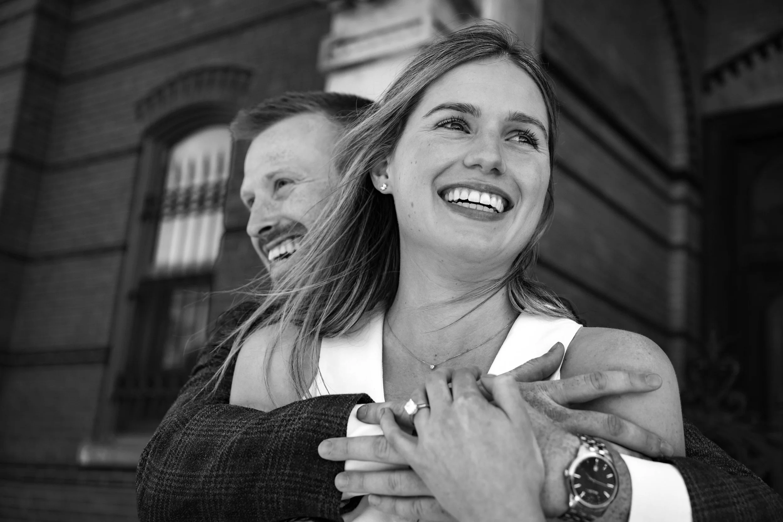 Black and white photo of a smiling woman and man hugging outdoors in front of a brick building.