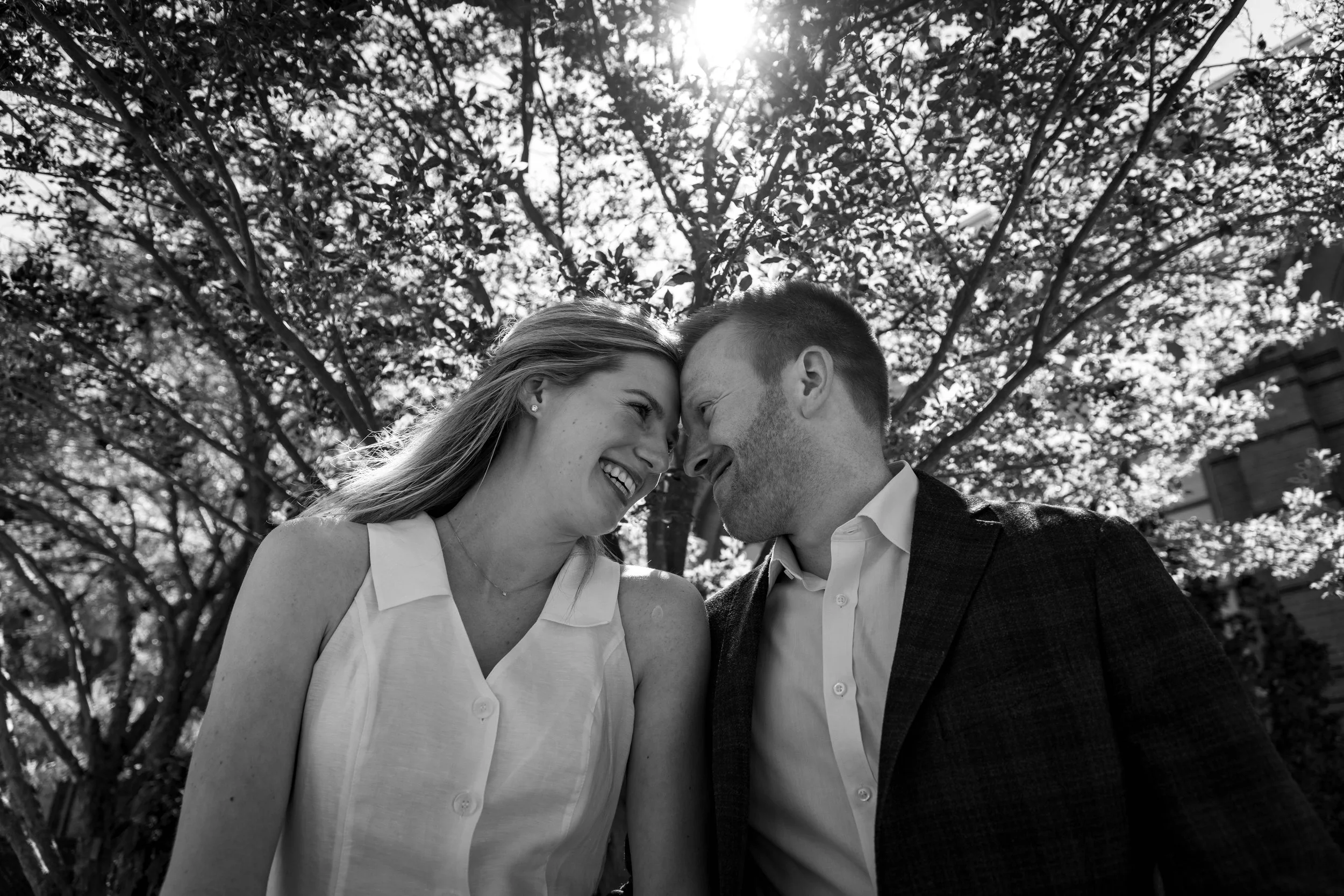 Black and white photo of a smiling couple with foreheads touching, outdoors under a tree with sunlight filtering through the leaves.
