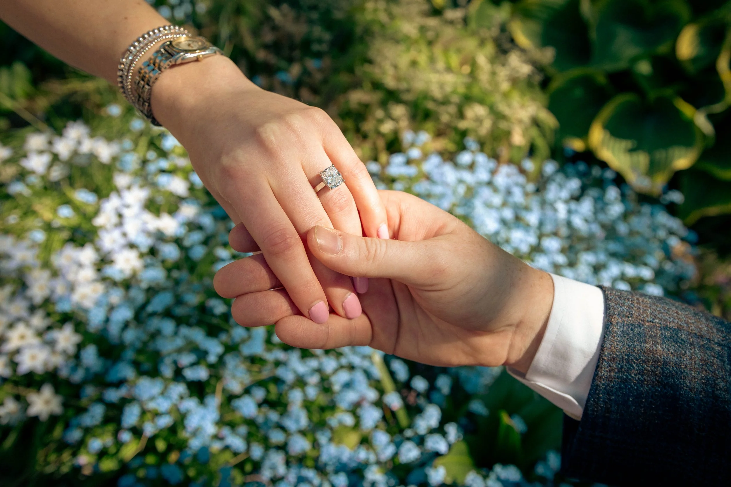 A person shows off an engagement ring on their finger while holding hands with another person outdoors, with trees and flowers in the background.