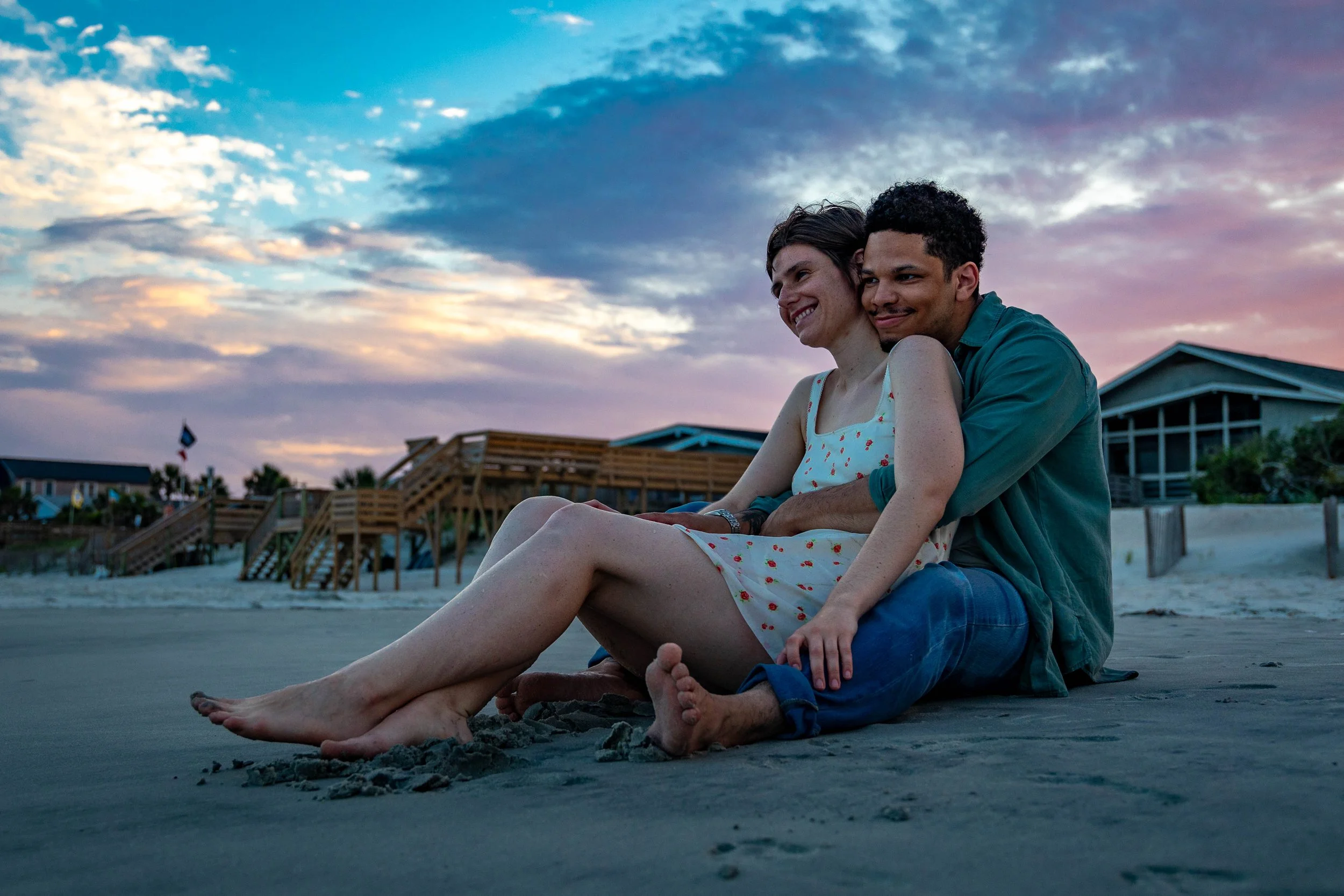 A couple sitting on the beach at sunset, smiling and enjoying each other's company, with houses and a wooden deck in the background.