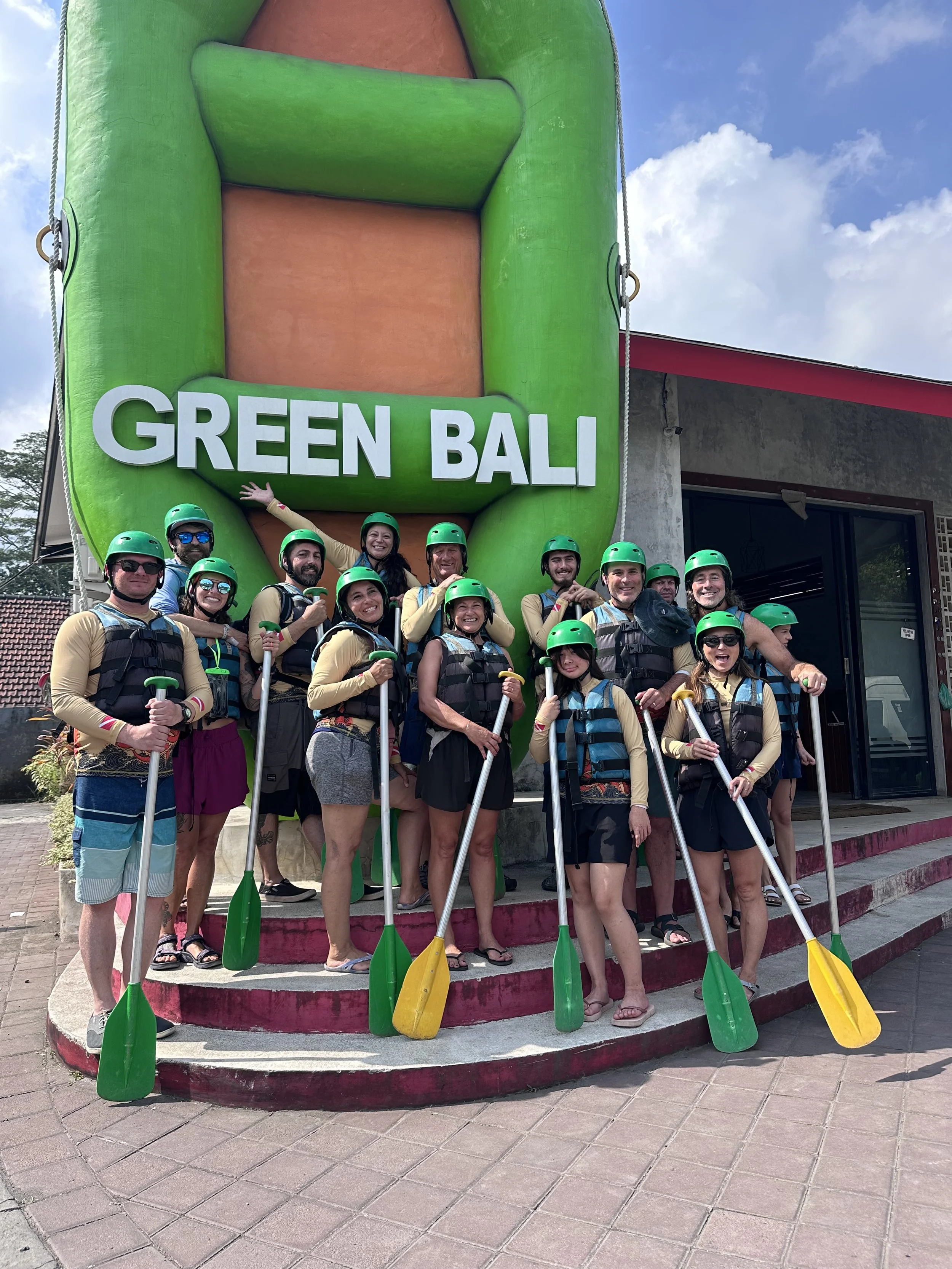 A group of people wearing green helmets and life vests, holding paddles, standing on steps in front of a large inflatable sign that says 'Green Bali'. Whitewater rafting adventure.
