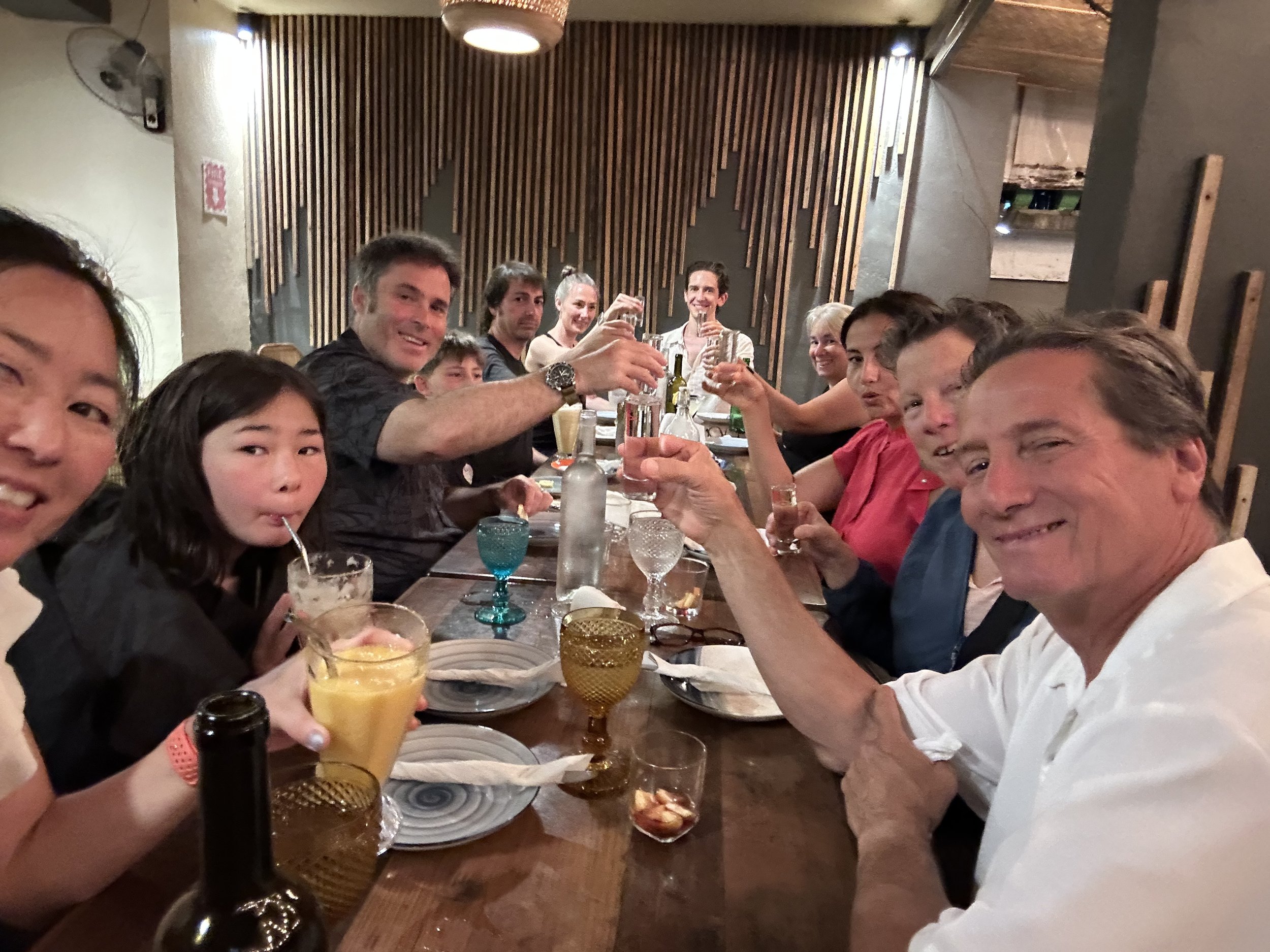 A group of people sitting around a long wooden table at a restaurant, enjoying drinks and smiling at the camera. The background features a modern wooden slat wall and ceiling light fixture.