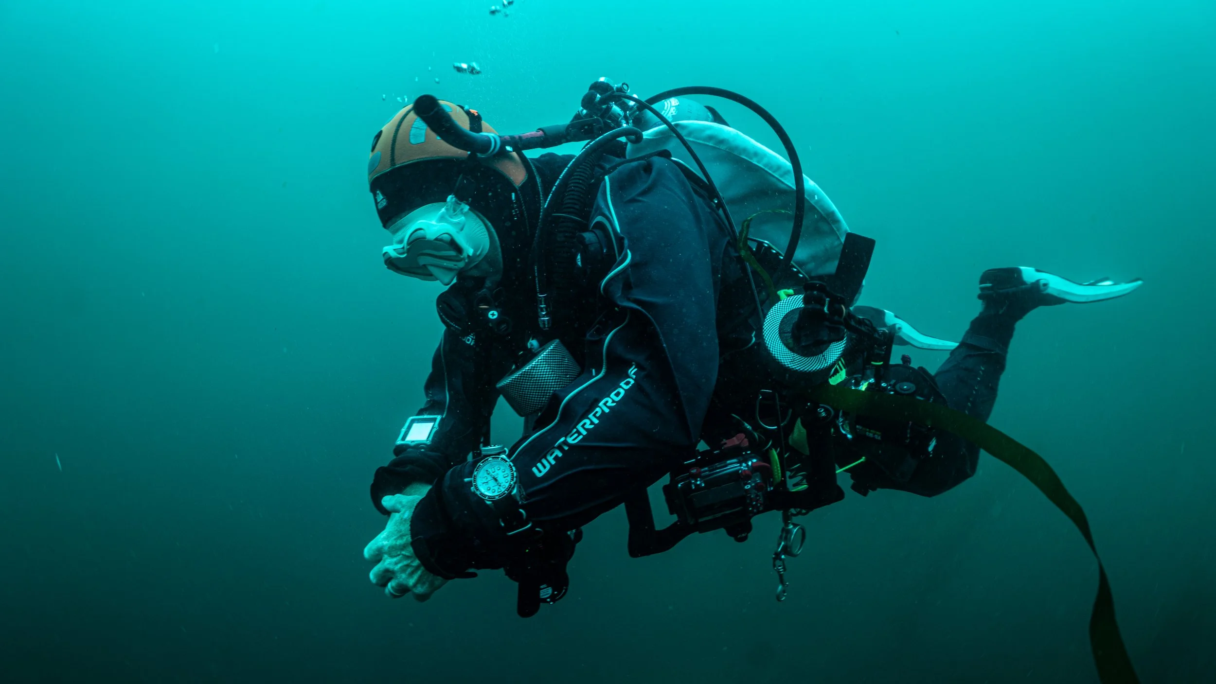A diver in full gear underwater looking at a smartphone or camera, surrounded by blue water.