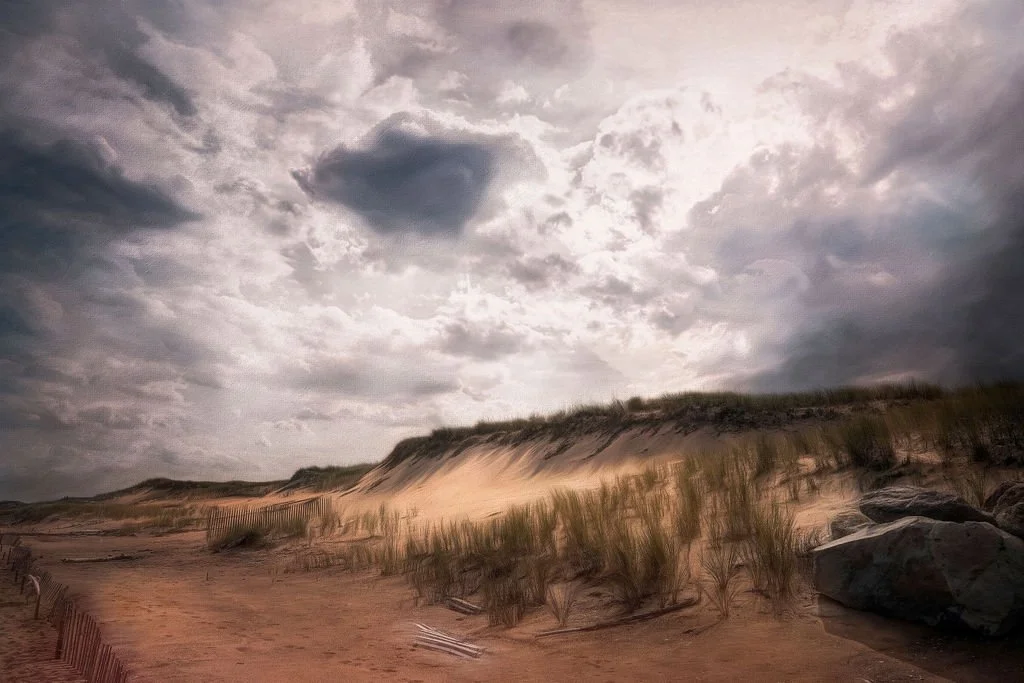 Sandy beach with dunes, grass, and rocks under a cloudy sky.