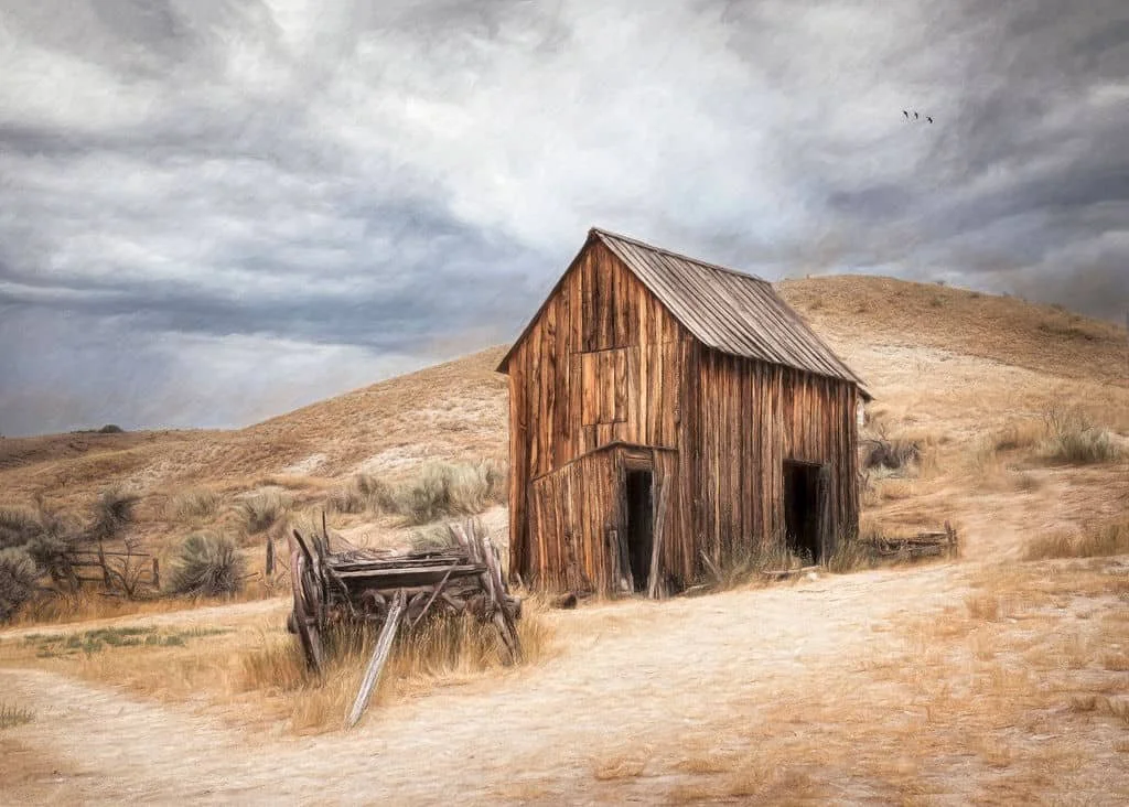 Rustic wooden barn in a western landscape with rolling hills, dry grass, and a dramatic cloudy sky in soft painterly tones.