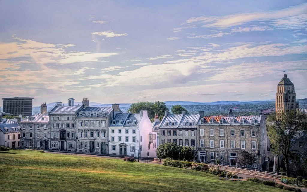 A row of historic buildings with gray and white facades under a partly cloudy sky, with a grassy park in the foreground and hills in the background.