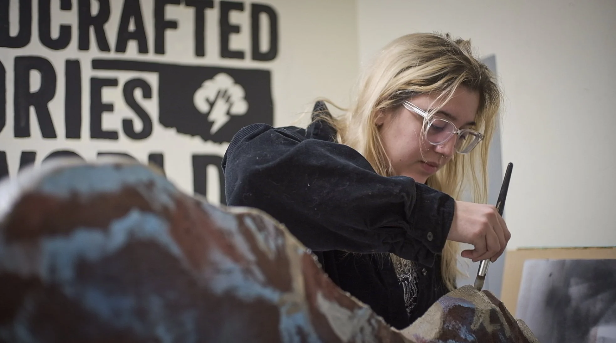 A young woman with blonde hair, glasses, and a septum piercing working on a large rock with a tool in an indoor setting with a painted sign on the wall.