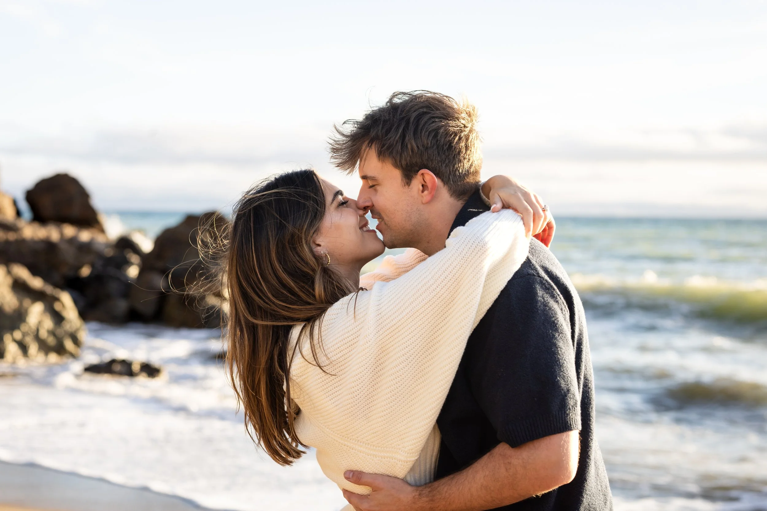 A couple sharing a moment on the beach, with the woman smiling and the man leaning in for a kiss, with waves and rocks in the background.
