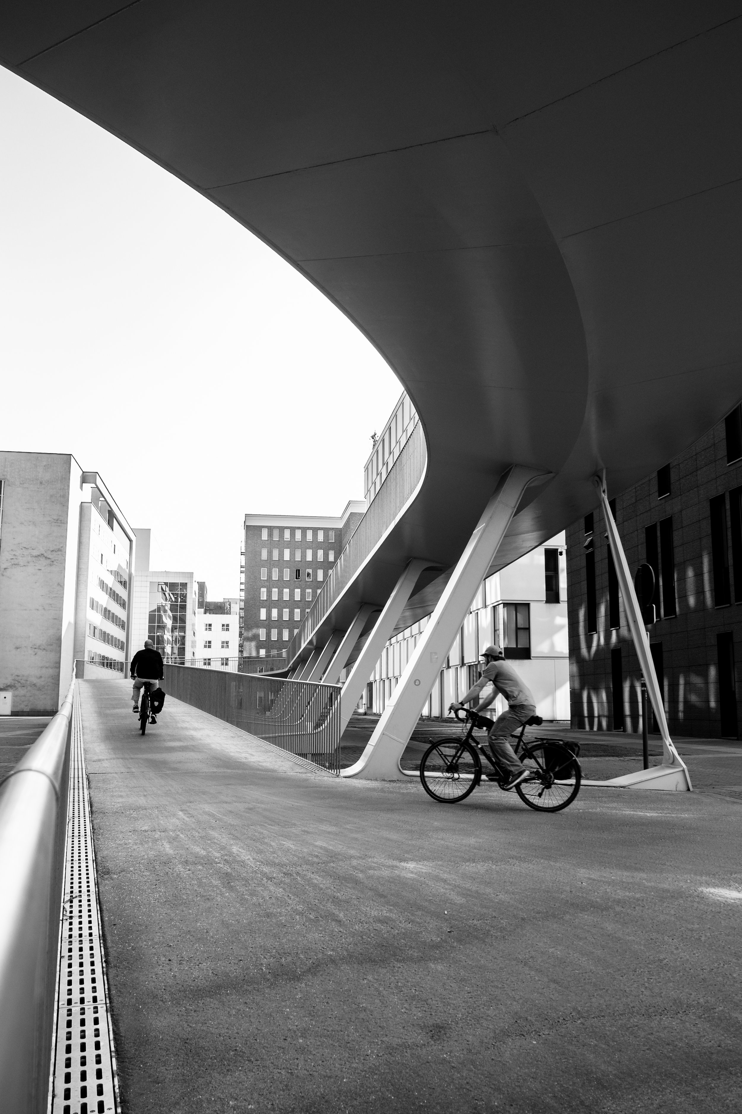 Twee mensen fietsen onder een moderne, gebogen architectural structuur in een stedelijke omgeving.