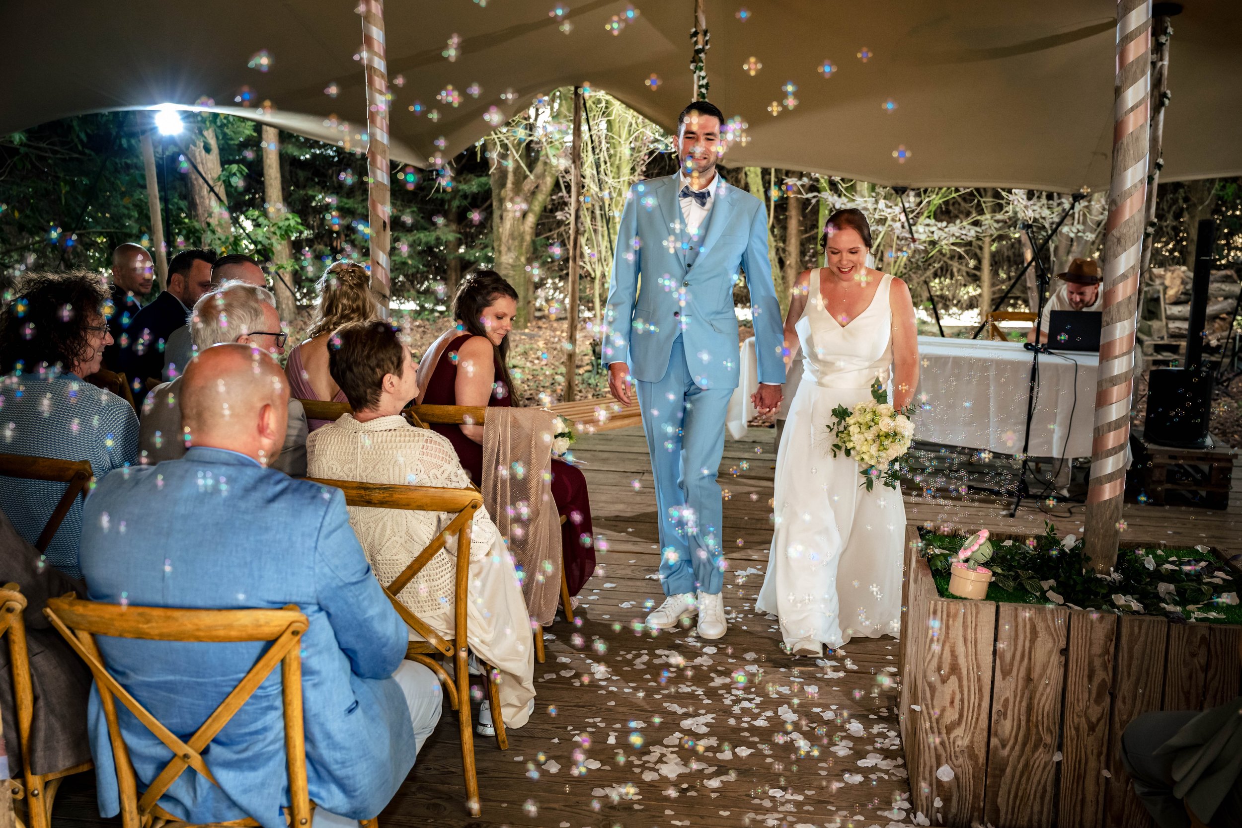Een bruiloft in de buitenlucht met een gekoppeld koppel dat hand in hand loopt, omringd door gasten die op een houten platform zitten. Er vallen confetti en ze lachen. De omgeving is bosachtig met veel groen en bomen.
