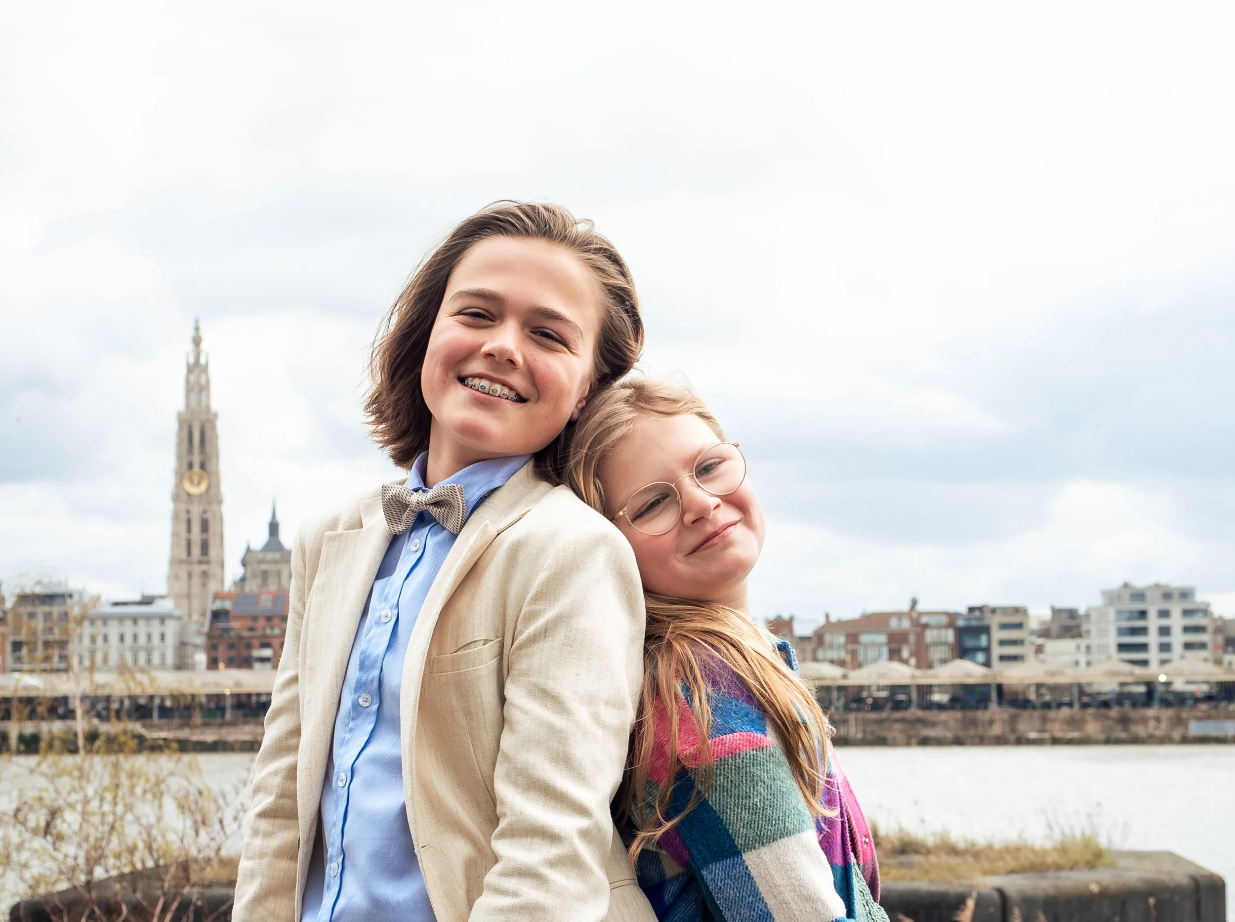 Twee lachende jonge meisjes staan dicht bij elkaar met de stad en een kerktoren op de achtergrond, onder een bewolkte hemel.