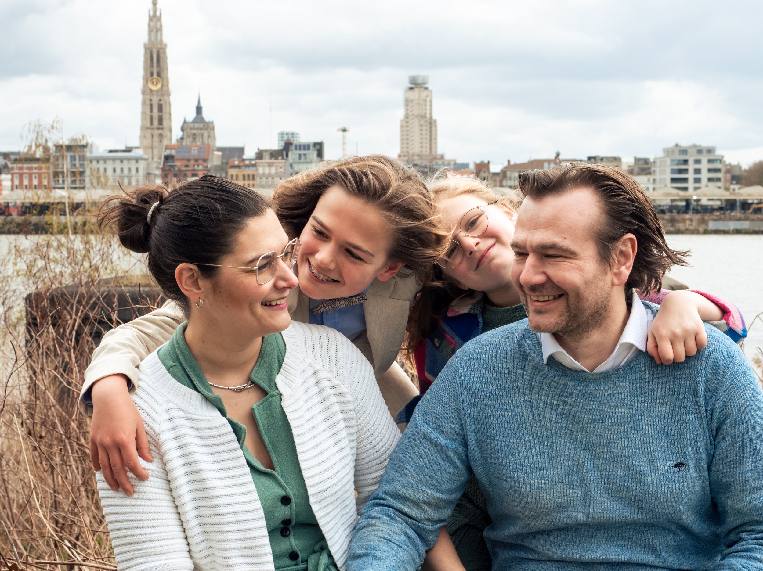 Vijf mensen die samen lachen en knuffelen bij een rivier met een stad op de achtergrond.