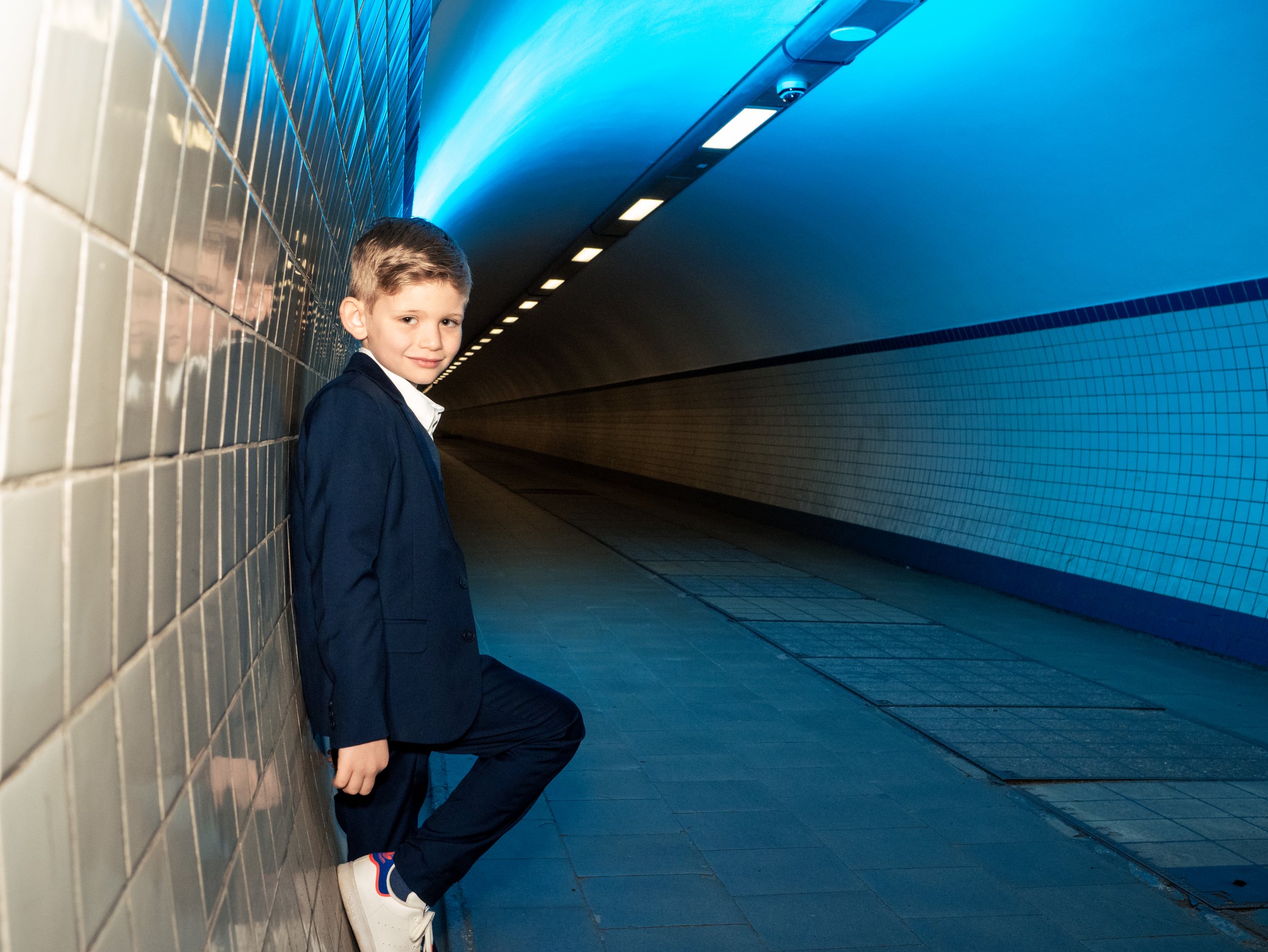 Klein jongetje in een donkerblauw pak leunt tegen de witte tegelwand in een ondergrondse tunnel, met een blauwe gloed op het plafond.