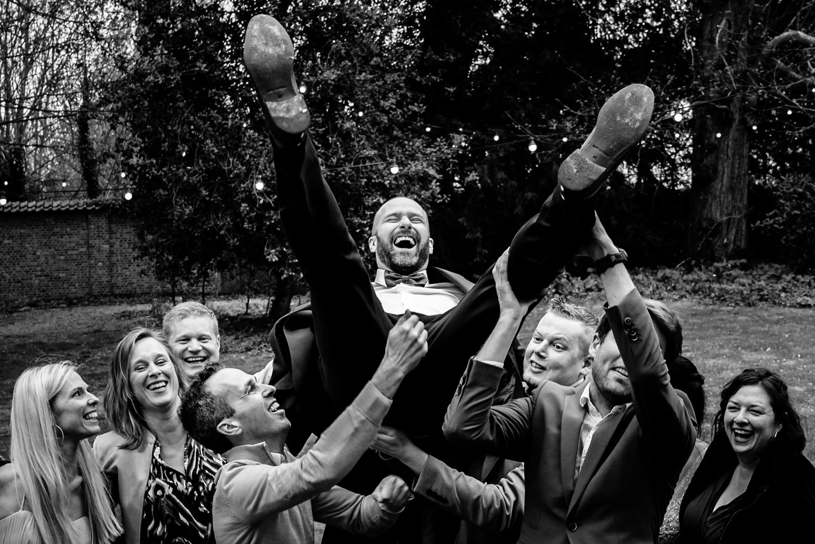 Een groep mensen in formele kleding die een man omhoog tilt, lachend en plezier hebbend op een buitenlocatie, zwart-wit foto.