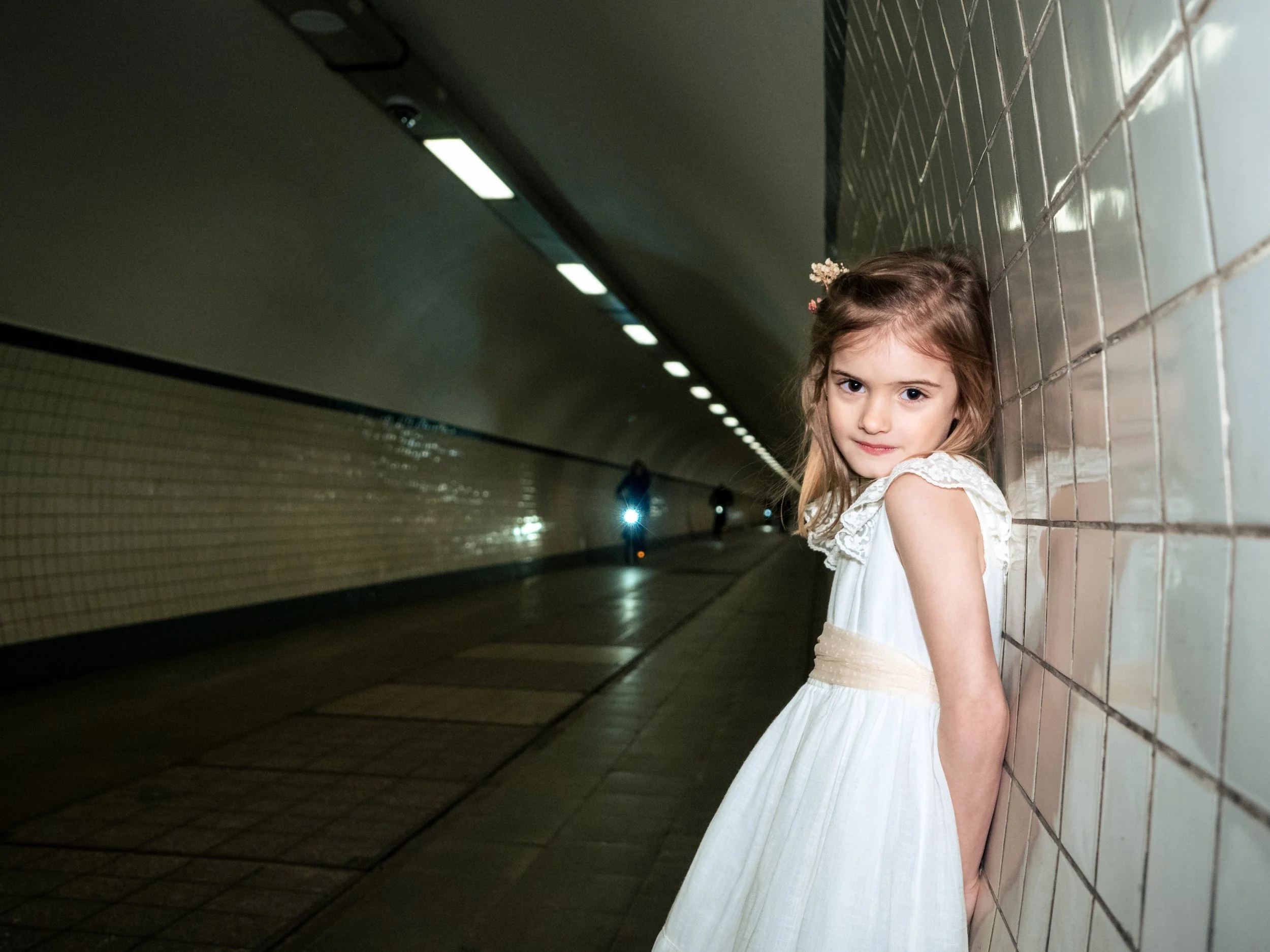Lieve jonge meisje in witte jurk leunt tegen een tegelmuur in een ondergrondse tunnel, met een zachte glimlach op haar gezicht.