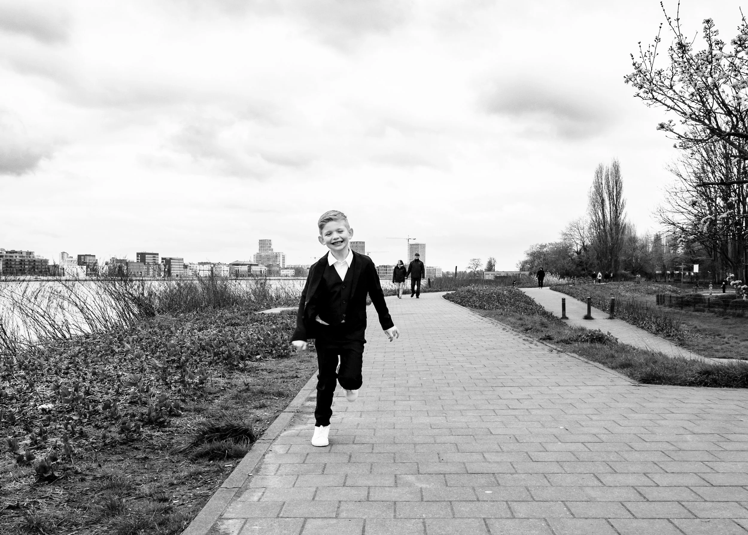 Een lachende jongen in een zwart pak rent op een wandelpad langs een stad samen met andere wandelaars op een bewolkte dag.