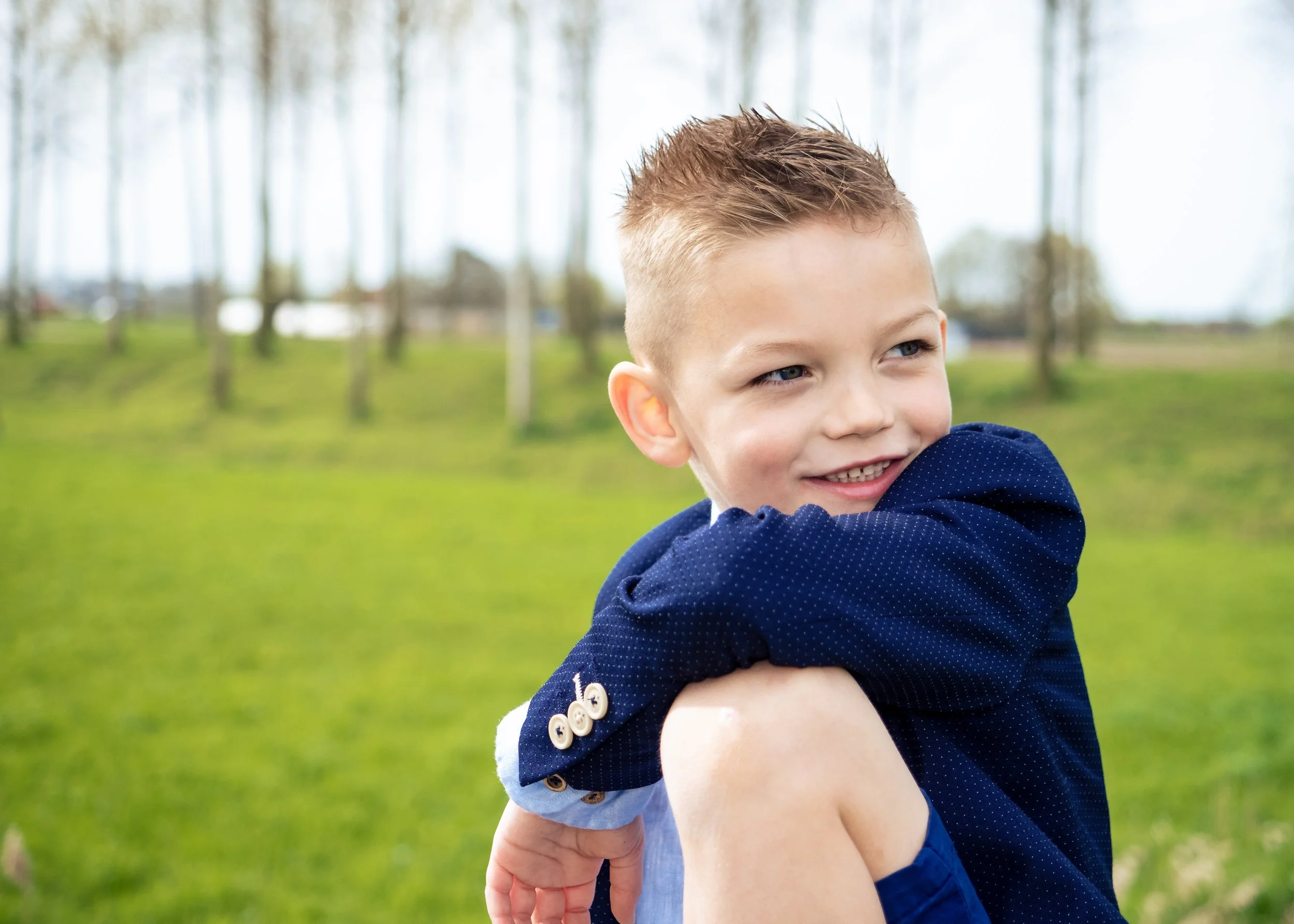 Lachende jonge jongen in een blauwe blazer en korte broek zittend op grasveld in een park, met bomen op de achtergrond.