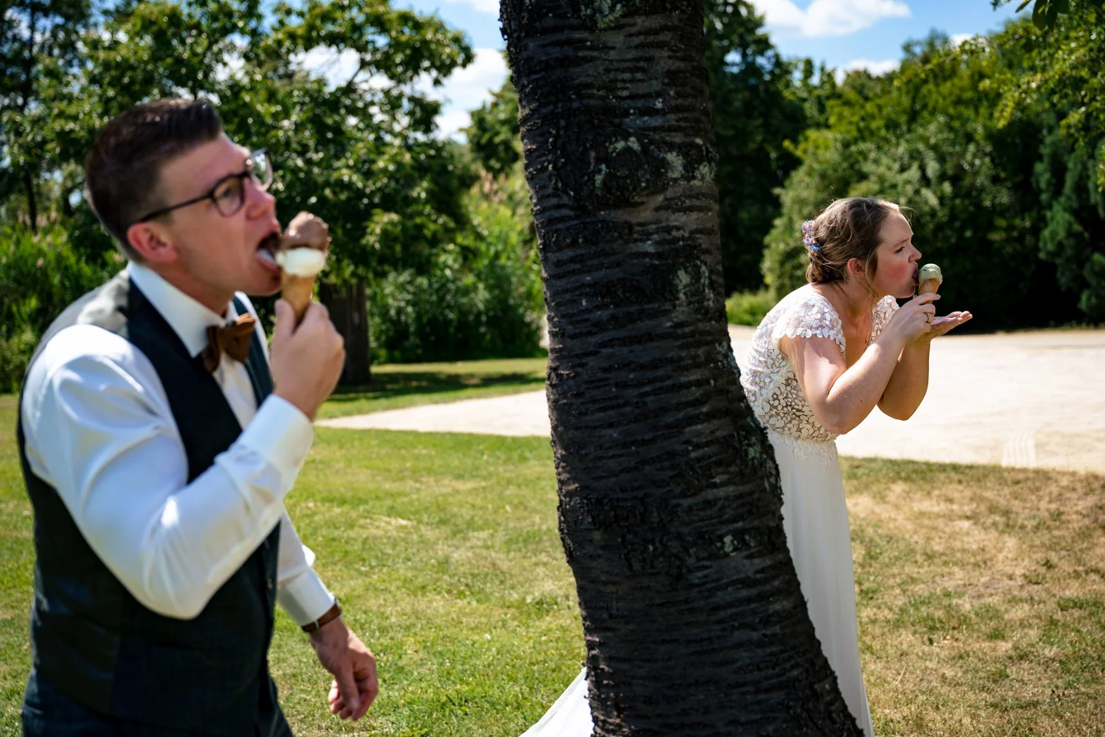 Een man en een vrouw, beiden in formele kleding, plezier makend terwijl ze ijs eten in een park met bomen, waarvan de vrouw achter een boom schuilt.