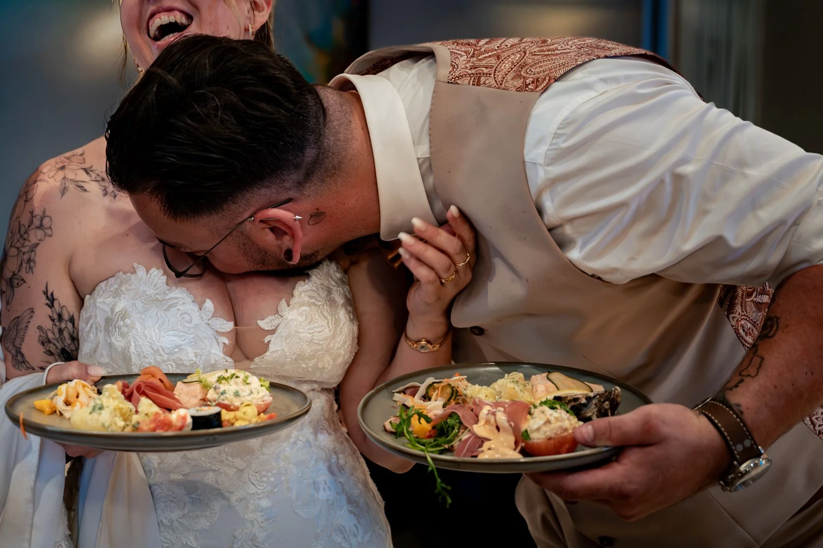Man en vrouw op bruiloft, man kust bruid, beiden lachen, ze houden platen met eten, bruid draagt witte jurk met kant, man draagt beige shirt met patroon op de mouw, ze hebben tatoeages, ze dragen sieraden, er is een feestelijke sfeer.