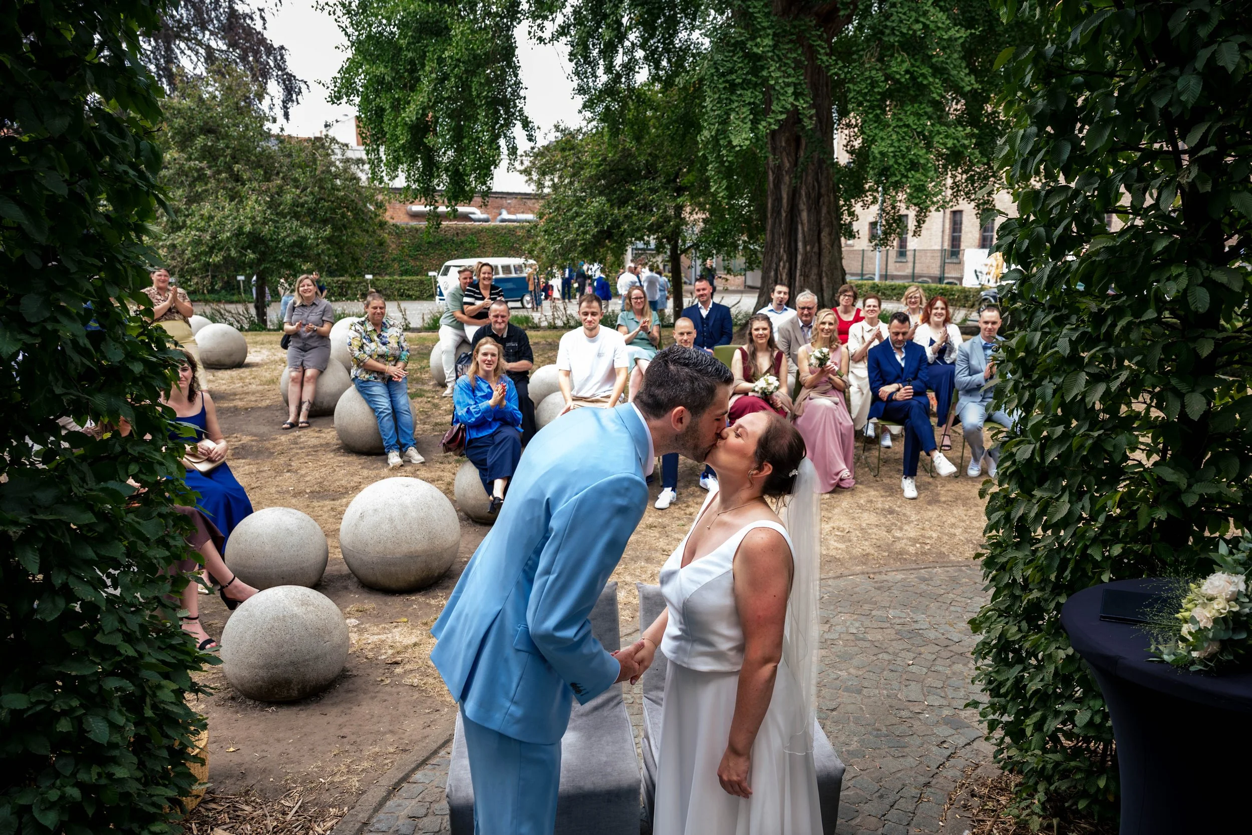 Een bruiloft buitenshuis met een bruid en bruidegom die elkaar kussen en hand in hand staan, omringd door gasten die kijken en applaudisseren, met groene bomen en een witte bestelbus op de achtergrond.