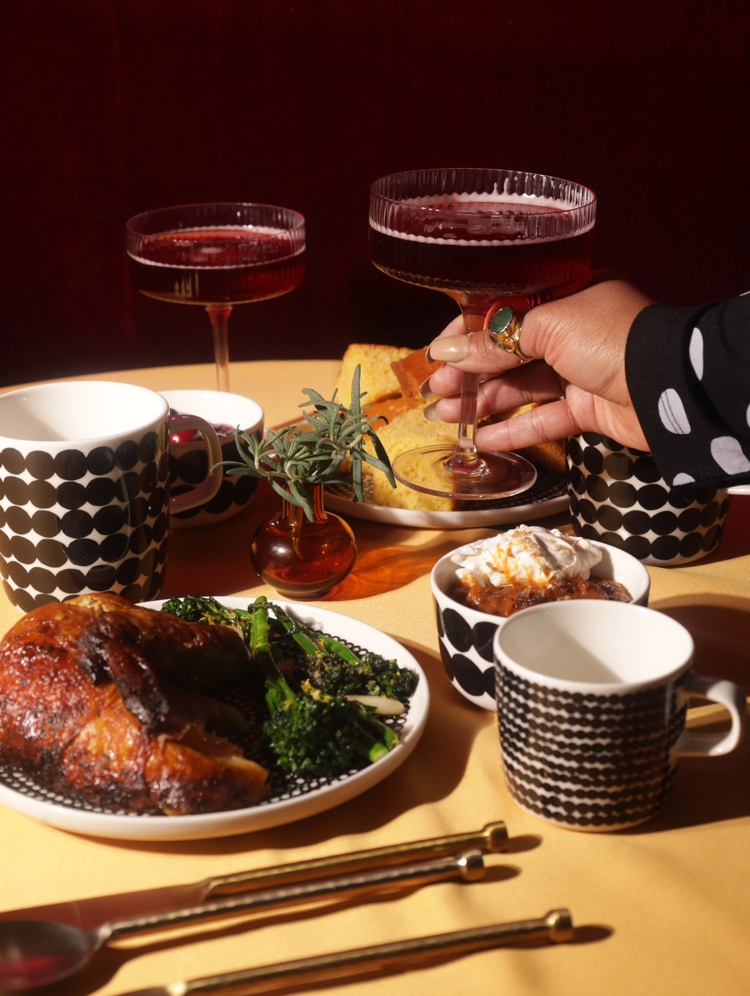 A dinner table with food and drinks, including two glasses of red wine, a mug and a cup with black and white patterns, roasted chicken with greens, bread, and dessert, with a small plant in a small amber vase and sunlight illuminating the scene.