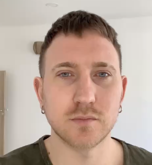 Close-up of a young man with short brown hair, blue eyes, and earrings, looking directly at the camera indoors.