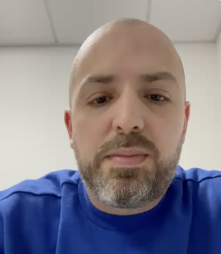 Close-up of a man with a shaved head and beard, wearing a blue shirt, in an indoor setting with a plain ceiling and wall.