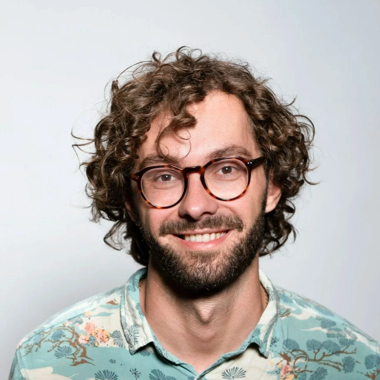 A smiling man with curly hair and glasses, wearing a floral patterned shirt, against a plain light background.