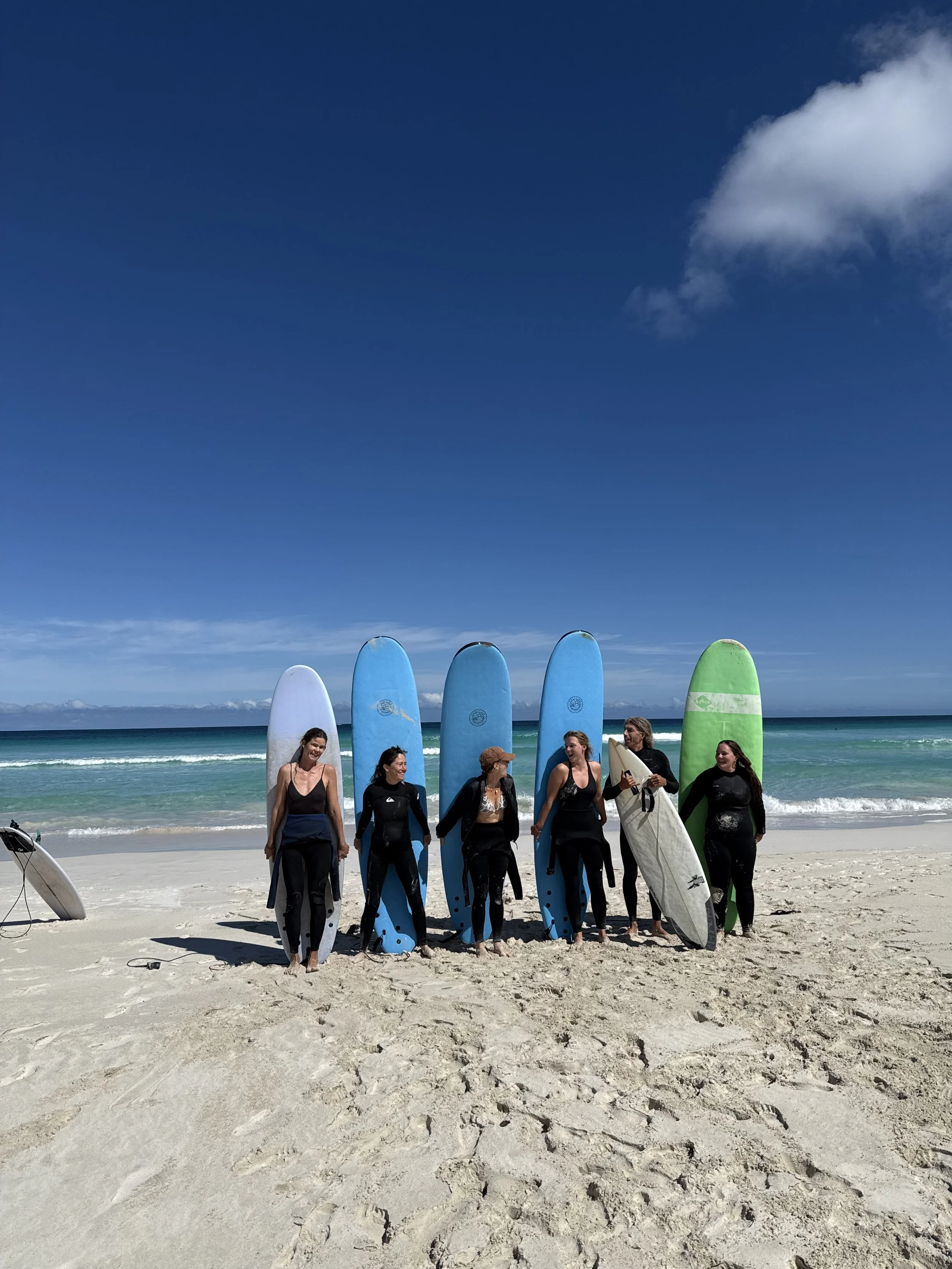 Group of six people standing on a beach holding surfboards, with the ocean and blue sky in the background.