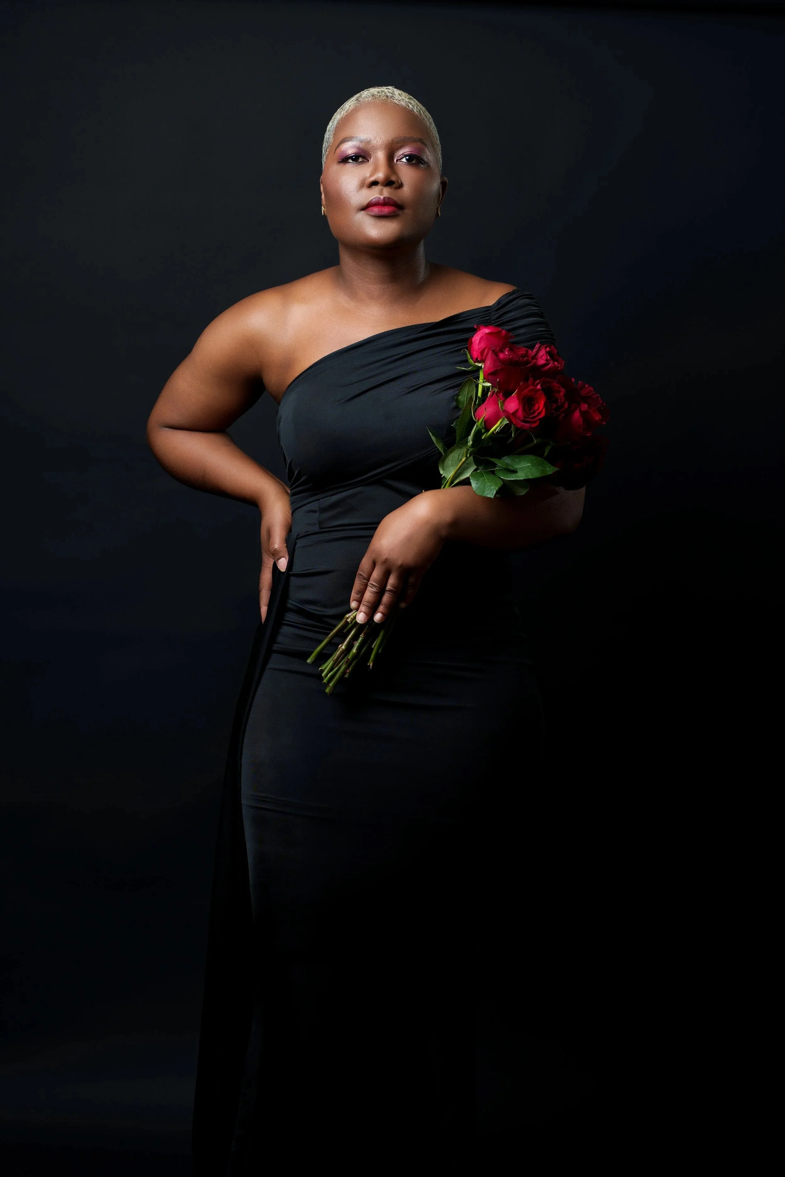 A woman in a black one-shoulder gown holding a bouquet of red roses against a dark background.