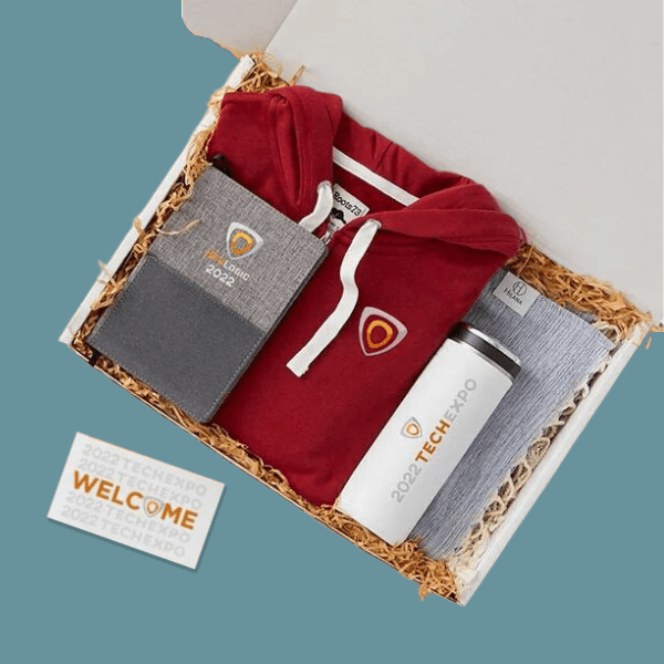 Gift box containing a red hoodie with logo, a gray notebook with logo, a white tumbler with logo, and a folded gray cloth, all arranged on shredded paper.