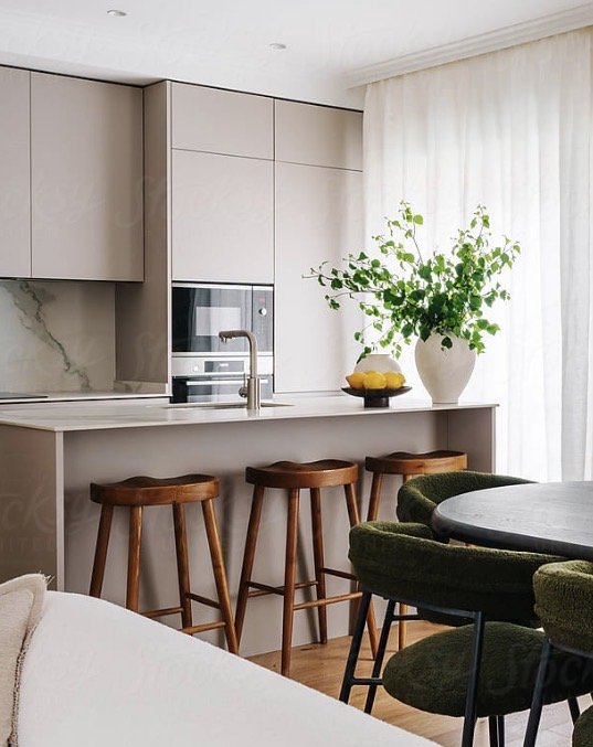 Modern kitchen with a white countertop island, wooden barstools, green upholstered chairs around a table, a large potted plant, and white curtains.