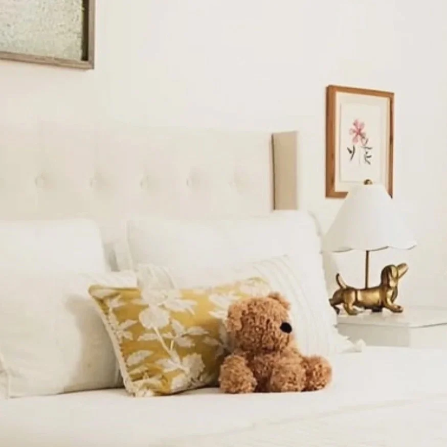 Bedroom project for an expat family in Rüschlikon along the lake of Zurich. A bedroom with a white bed, a teddy bear, patterned pillow, lamp with dachshund base, and framed floral artwork on the wall.