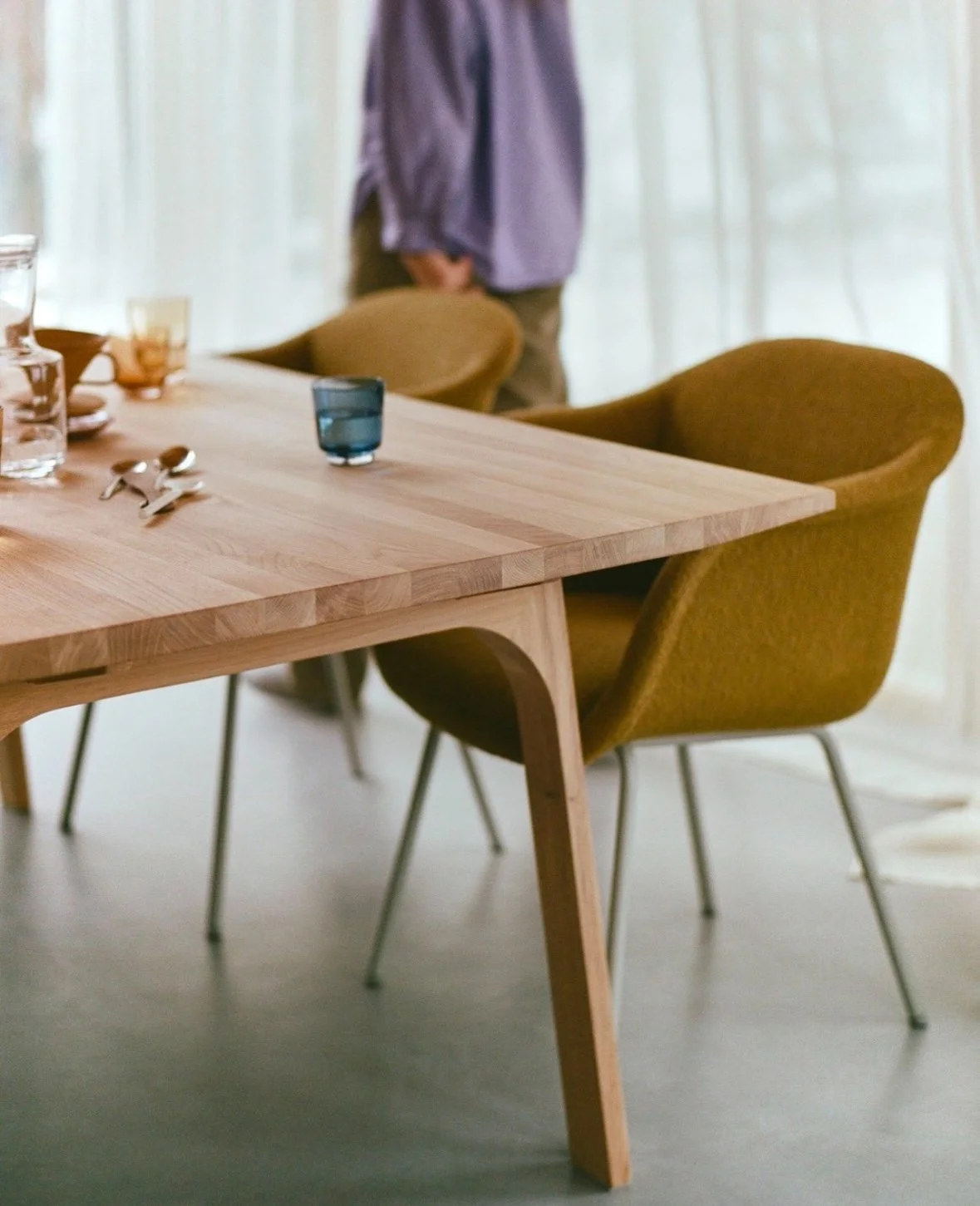 A wooden dining table with chairs, some glasses, and silverware, with a person standing in the background.