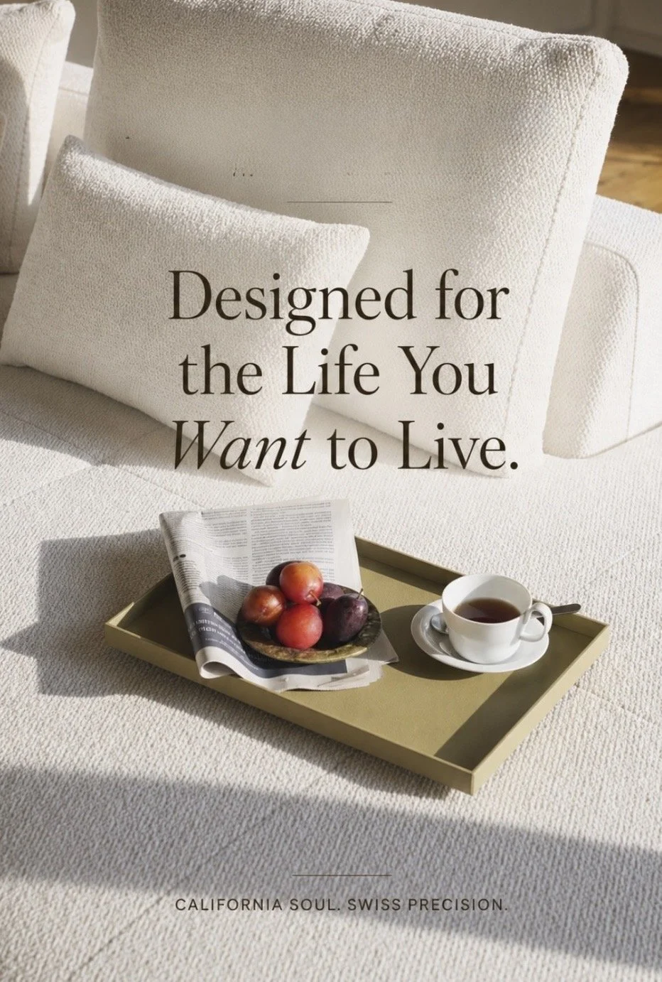 A cozy scene featuring a white cushioned chair with a matching pillow, a tray with apples, a newspaper, and a cup of tea on a light-colored textured surface. The overlay text reads "Designed for the Life You Want to Live" and at the bottom, "California Soul. Swiss Precision.".