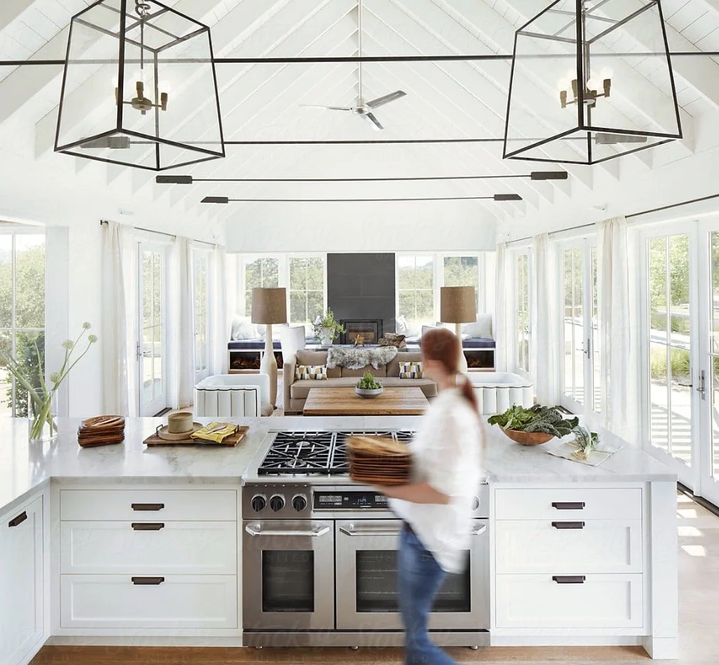 Bright open-concept kitchen and living room with white cabinetry, large windows, modern light fixtures, and a person carrying a plate of food.