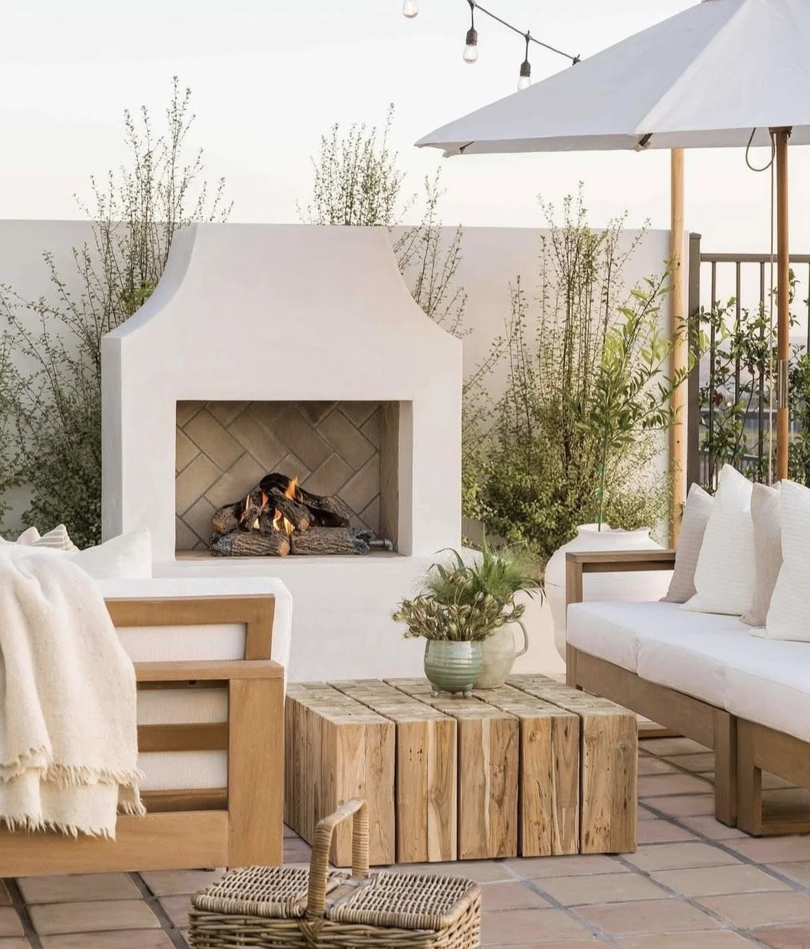 Outdoor patio with a white fireplace, white sofa with cushions, wooden table with a houseplant, an umbrella, and string lights at sunset.