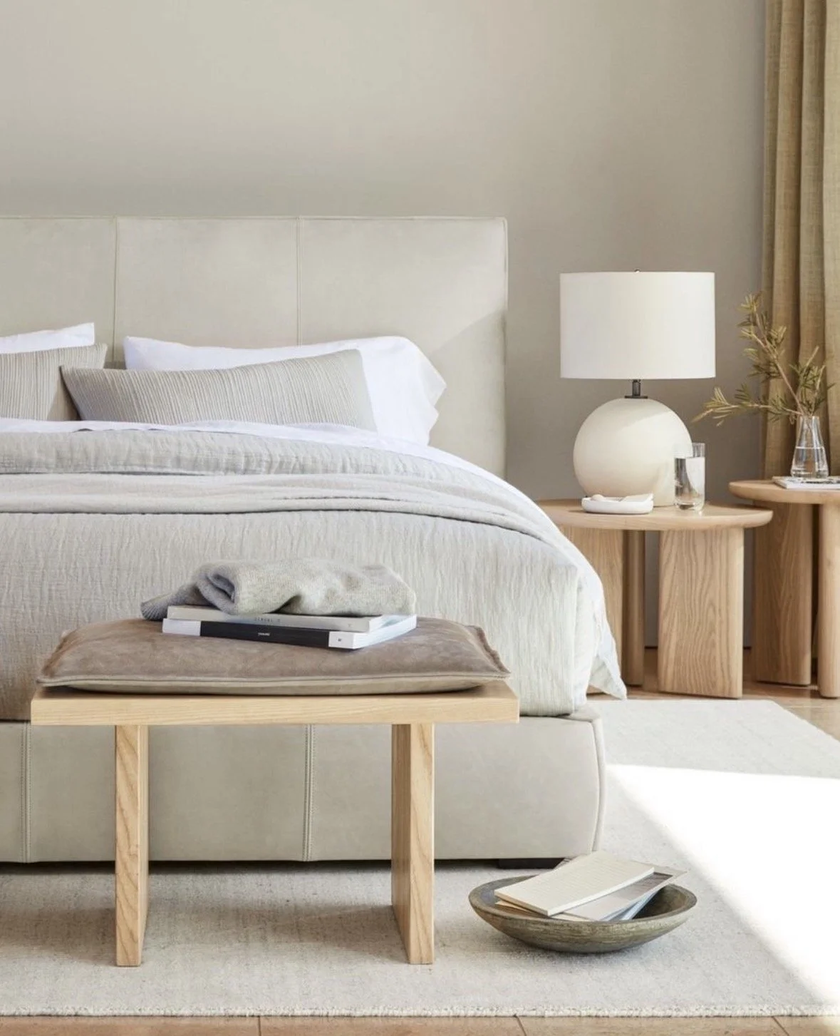 A neatly made bed with a beige upholstered headboard, white and beige pillows, and a cream-colored bedspread. Next to the bed are two round wooden side tables; one with a white lamp, a glass of water, and a small decorative object, the other with a t