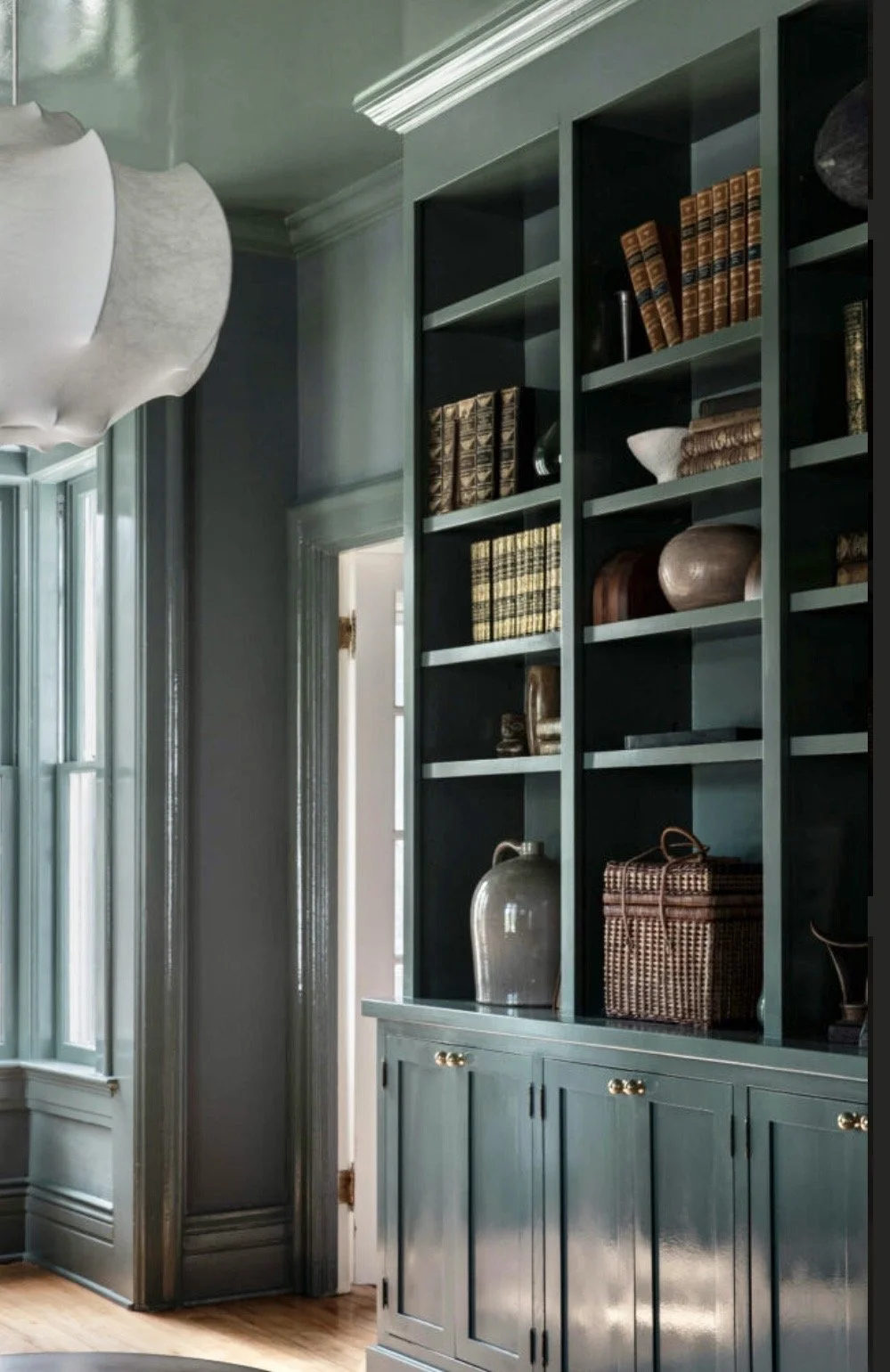 A large built-in bookshelf painted dark green filled with decorative books and pottery, with a large window and a white, cloud-shaped ceiling light fixture in a room with wooden flooring.