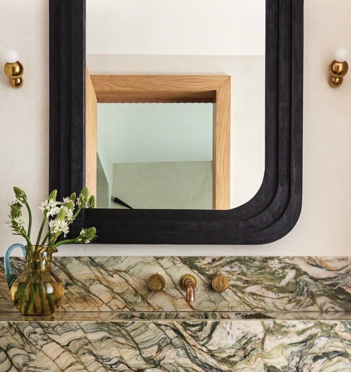 A bathroom vanity with a marbled countertop, a faucet, and a glass vase with white flowers. A mirror with a black and wood frame is above the vanity, reflecting a doorway and ceiling.