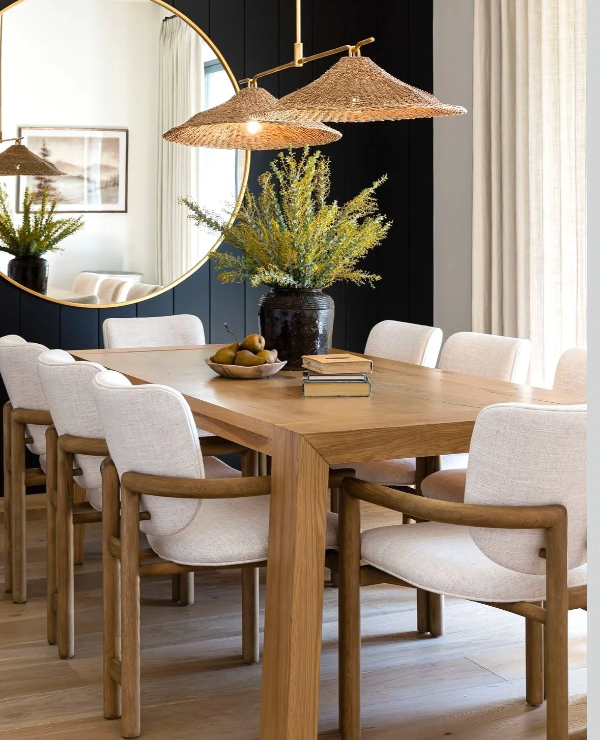 A dining room with a wooden table, surrounded by eight white upholstered chairs with wooden arms. A large black vase with yellow-green flowers sits on the table, along with a bowl of pears and three stacked books. A round mirror on a black wall refle